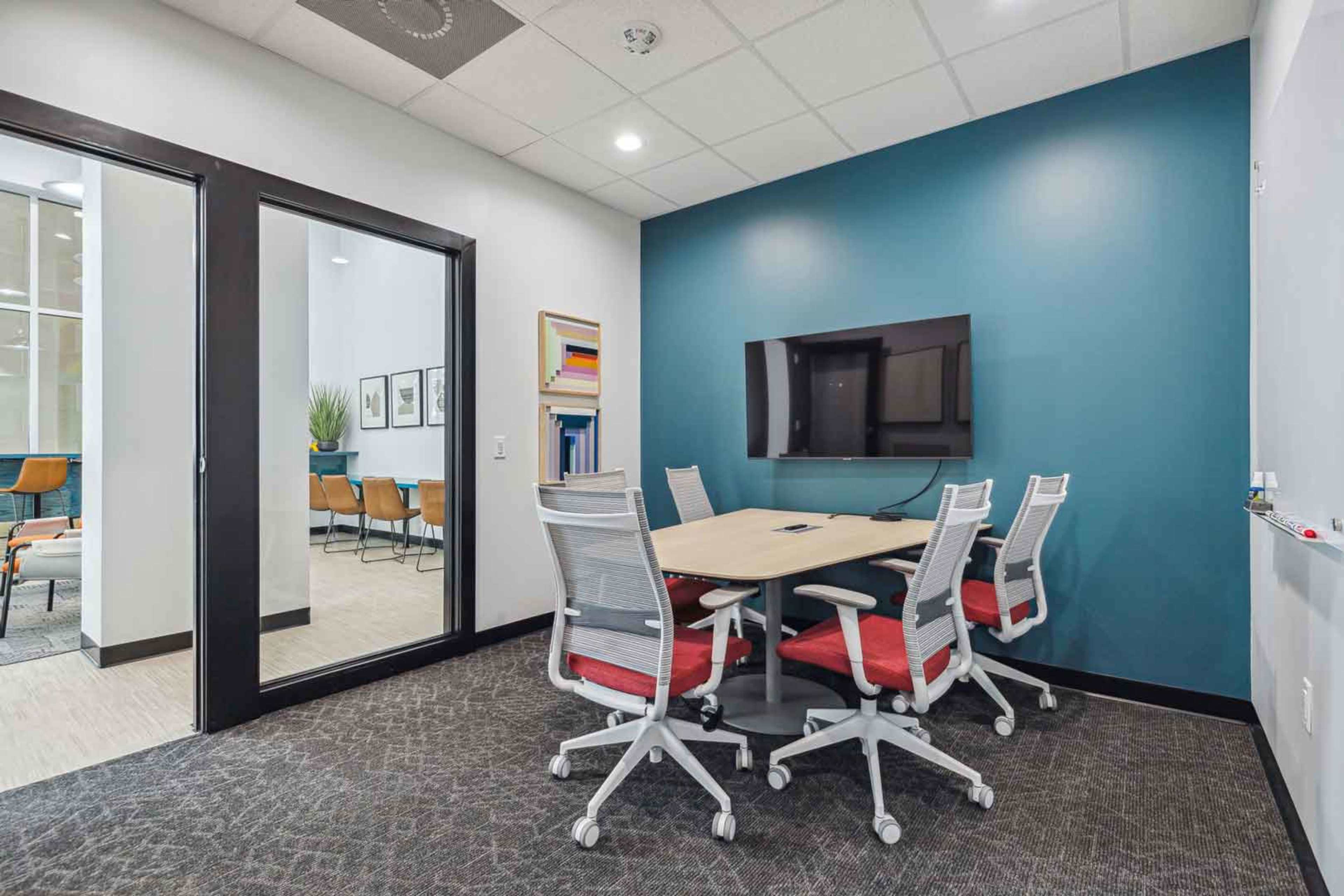 A small conference room features a round table surrounded by six ergonomic chairs and a wall-mounted TV.