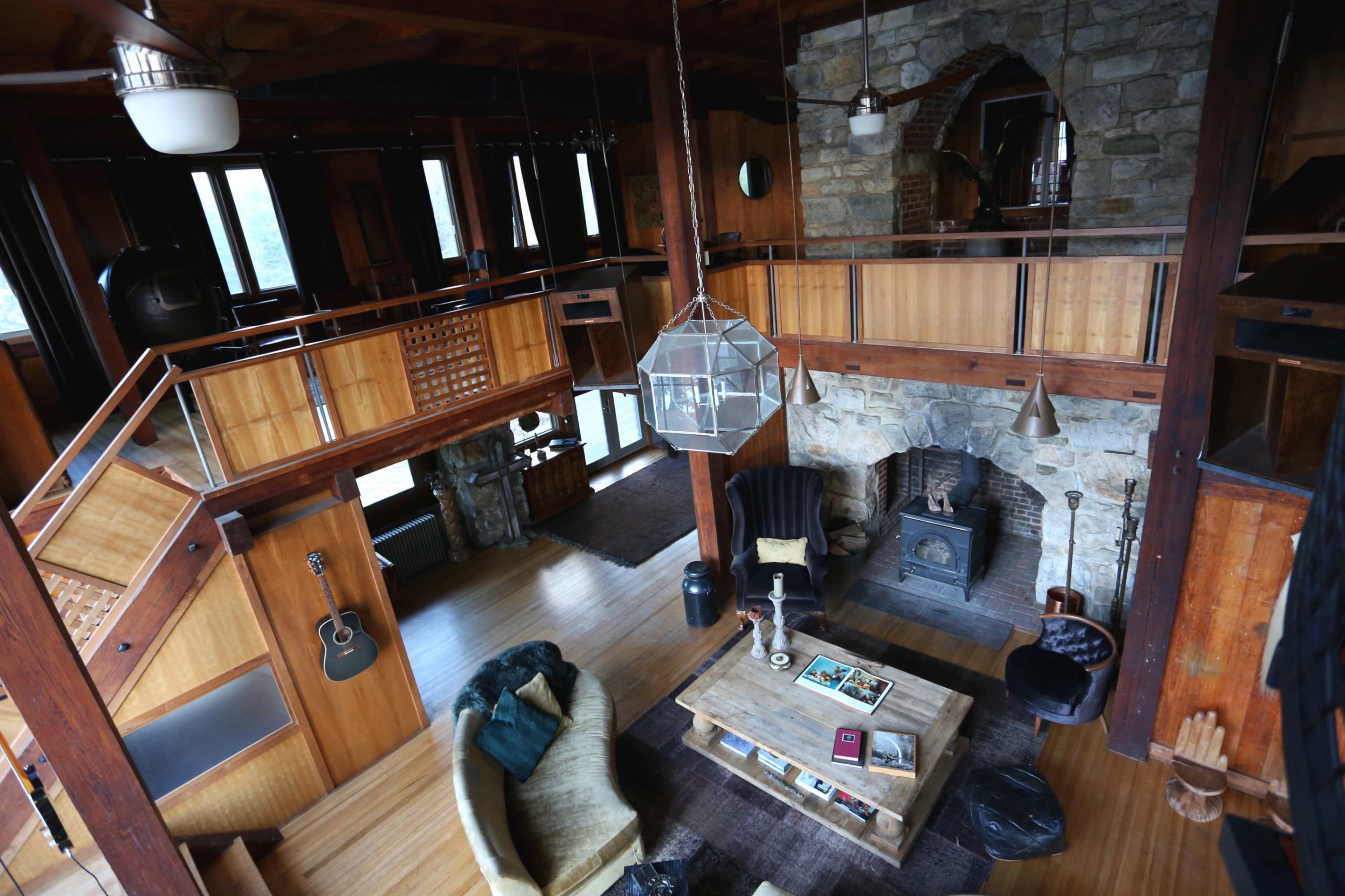 The interior of a modern wooden home features a stone fireplace, a central coffee table, and a balcony overlooking the living area.