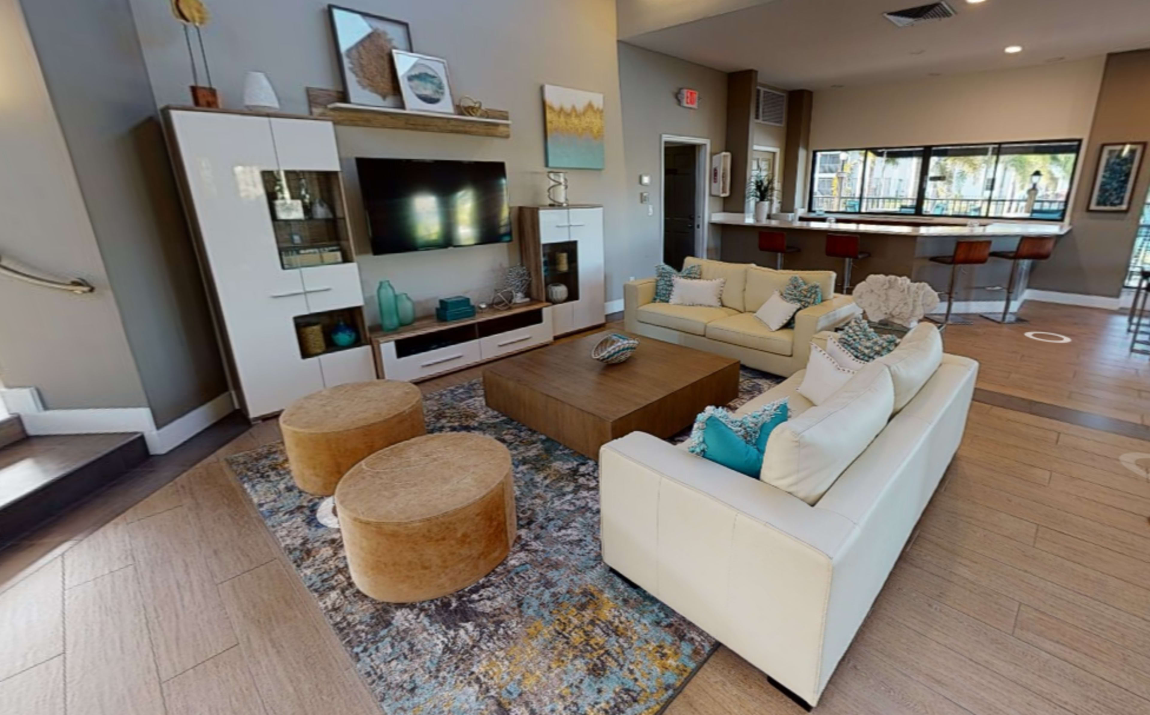 The image shows a modern living area with a white sofa, two round ottomans, a coffee table, and a television unit, all arranged around a patterned rug.