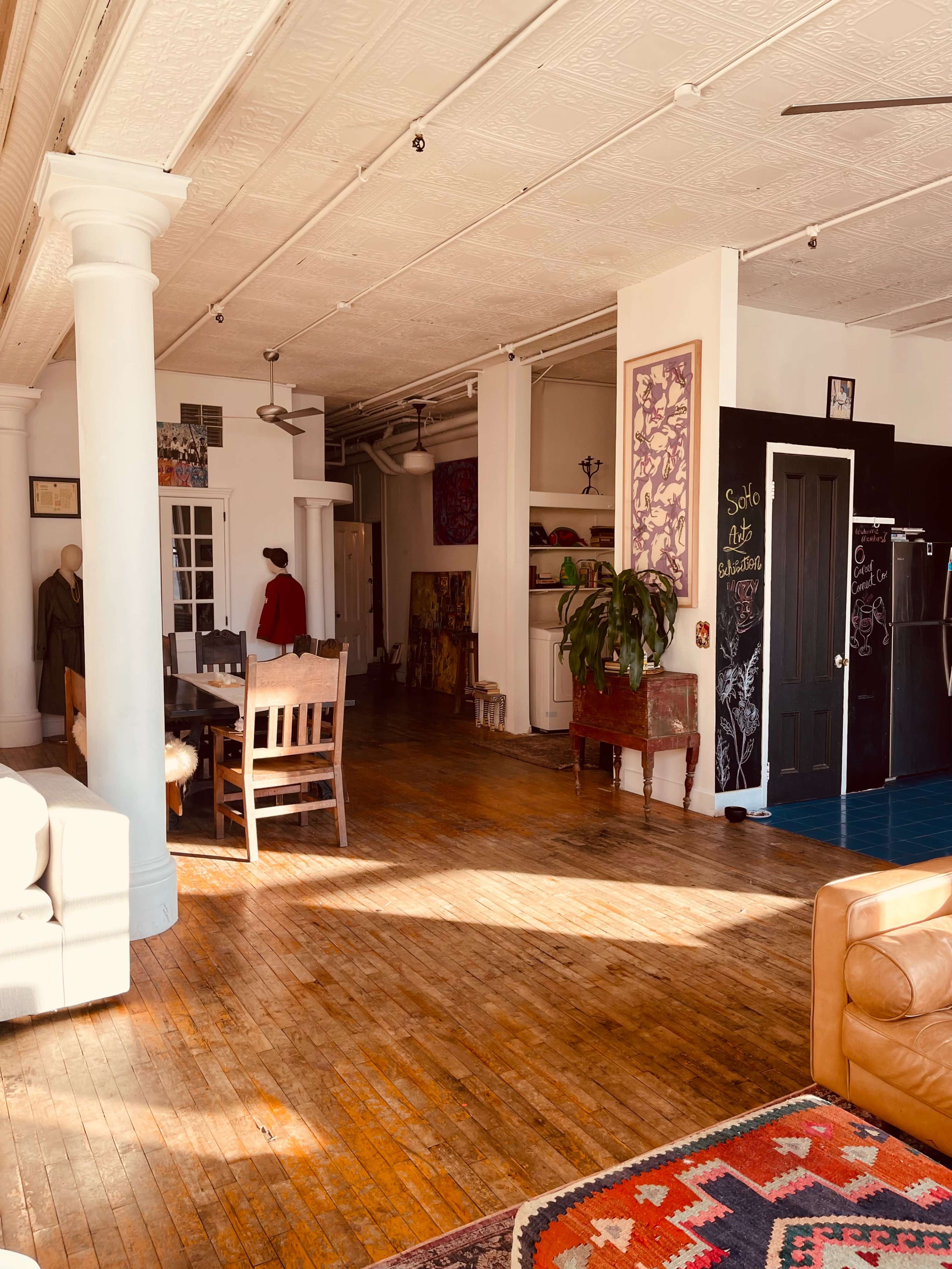 The image shows a spacious, well-lit interior of a room featuring wooden floors, a dining table with chairs, a couch, and art displayed on the walls.