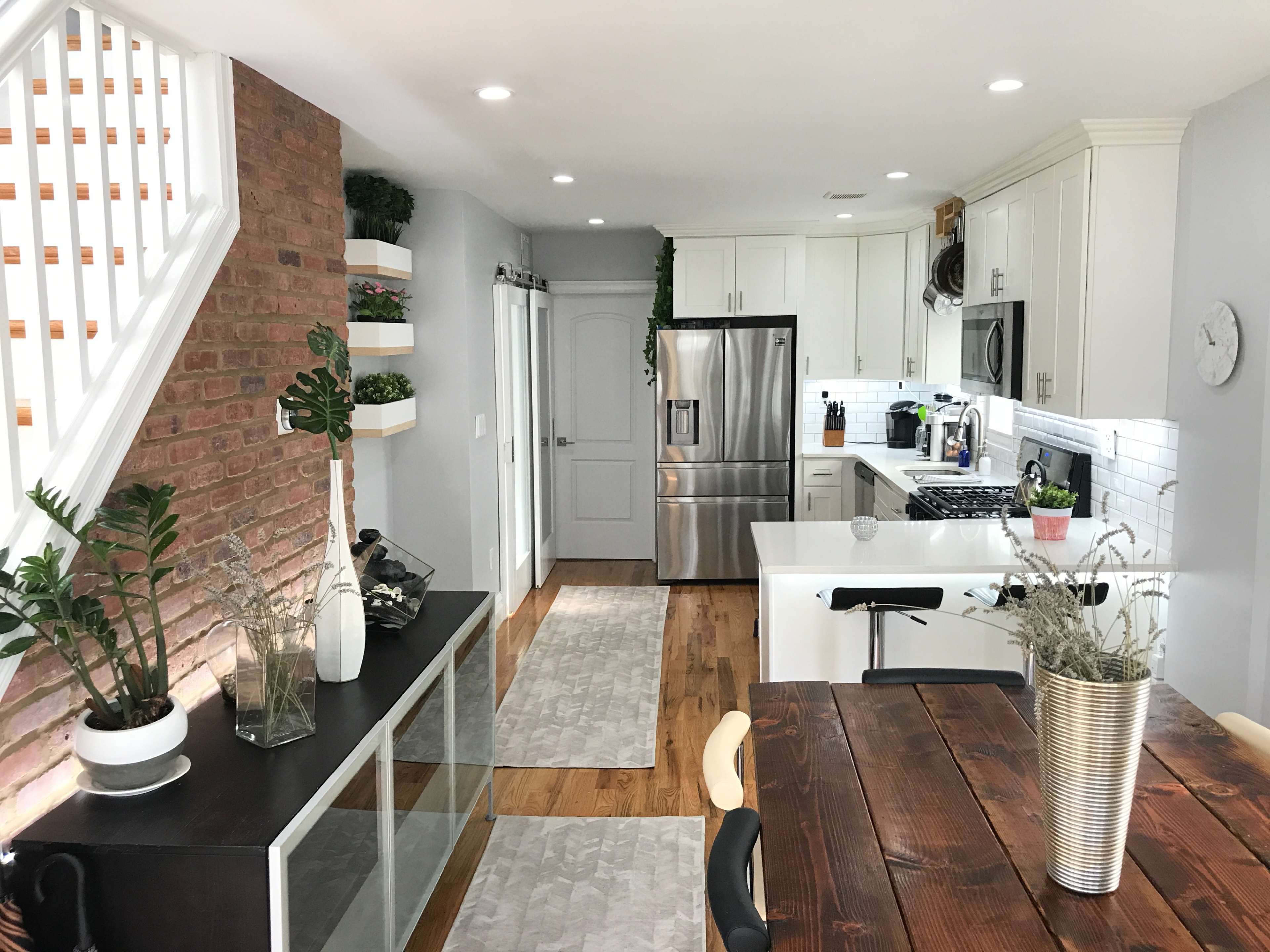 The image shows a modern kitchen and dining area featuring a brick accent wall, stainless steel appliances, and a wooden dining table.