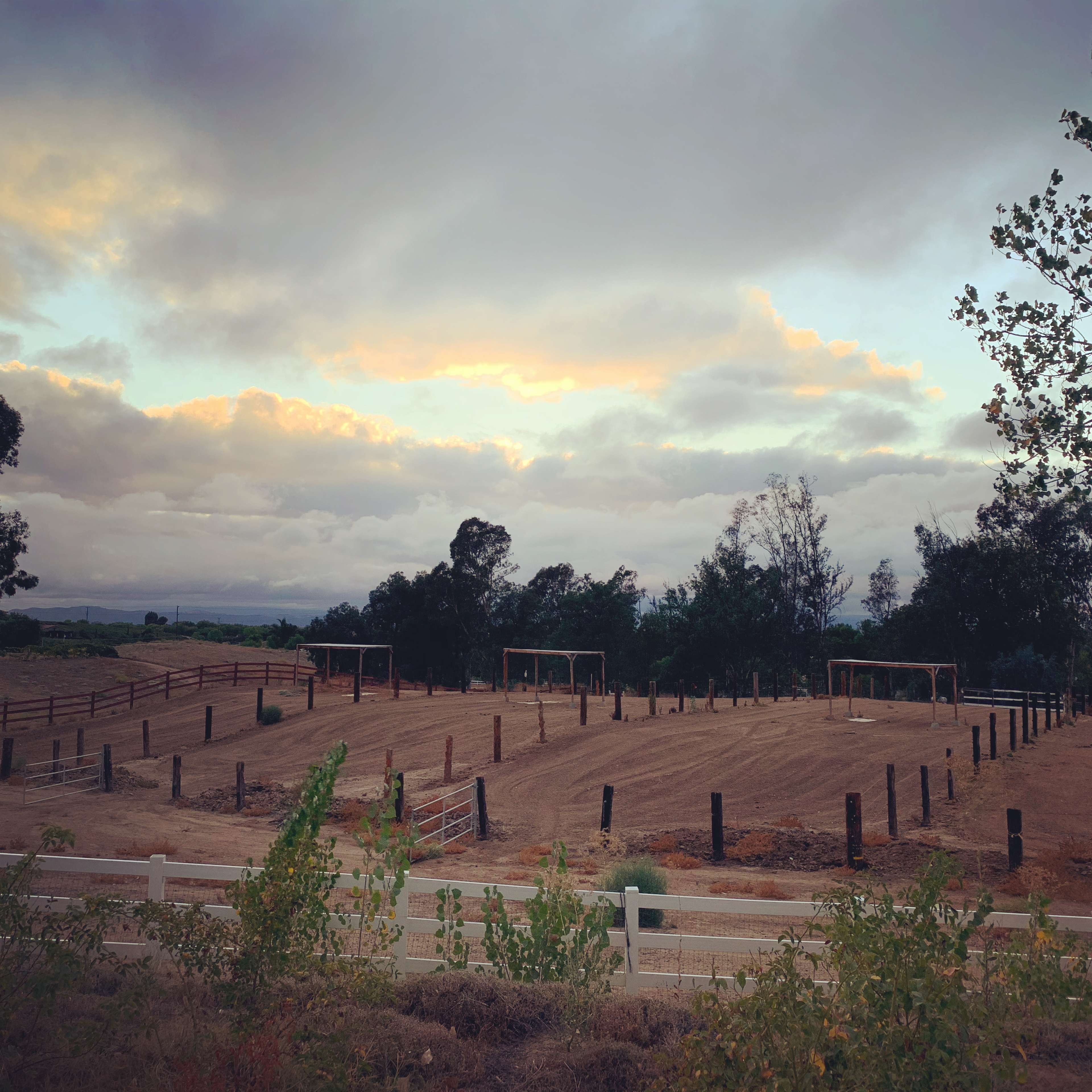 A fenced agricultural area with bare soil and several wooden structures is surrounded by trees under a cloudy sky.