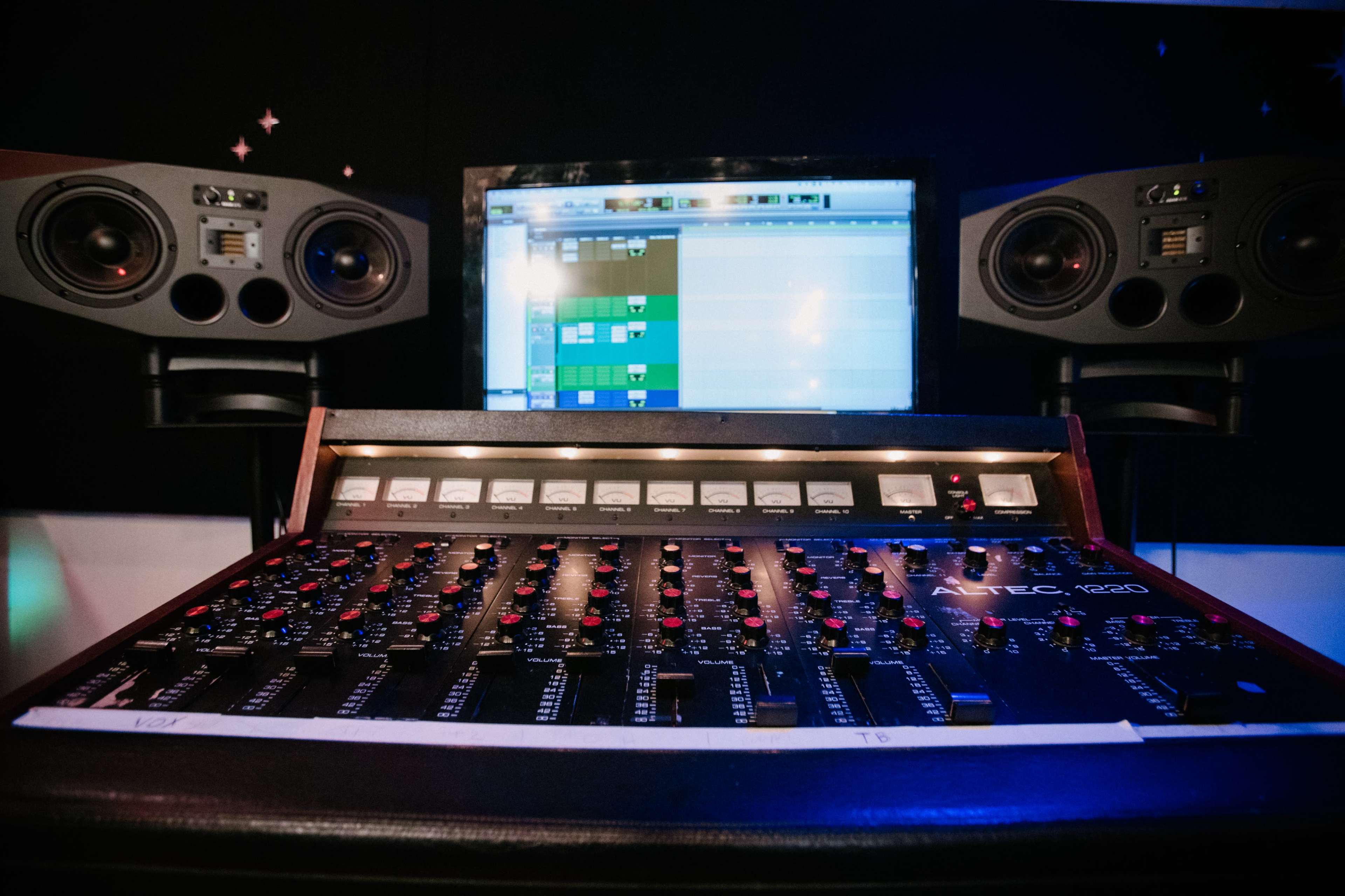 A mixing console with numerous controls is positioned in front of computer monitors displaying audio editing software and studio speakers.