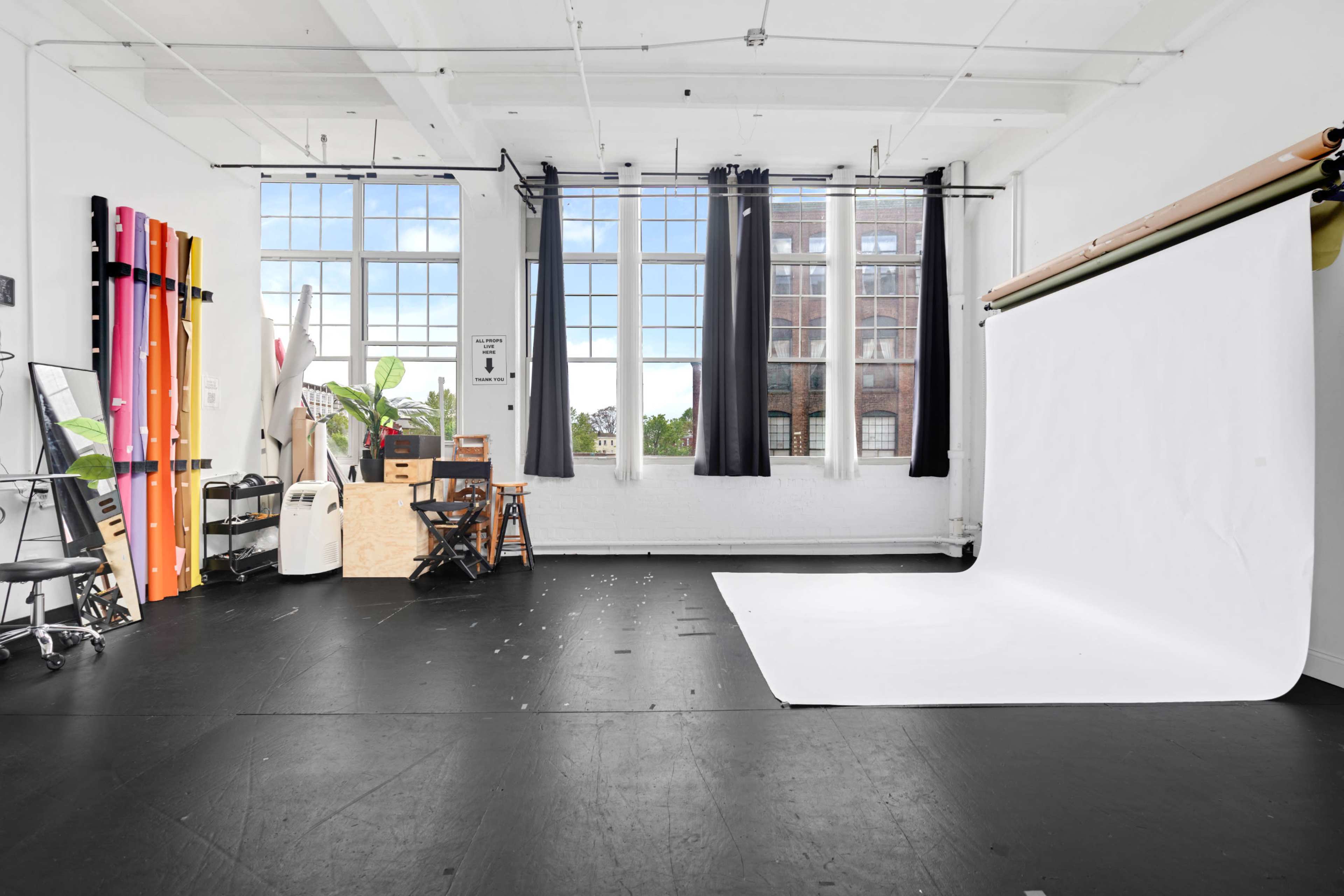 The image shows a spacious studio with large windows, a simple white backdrop, and various props and equipment scattered around.