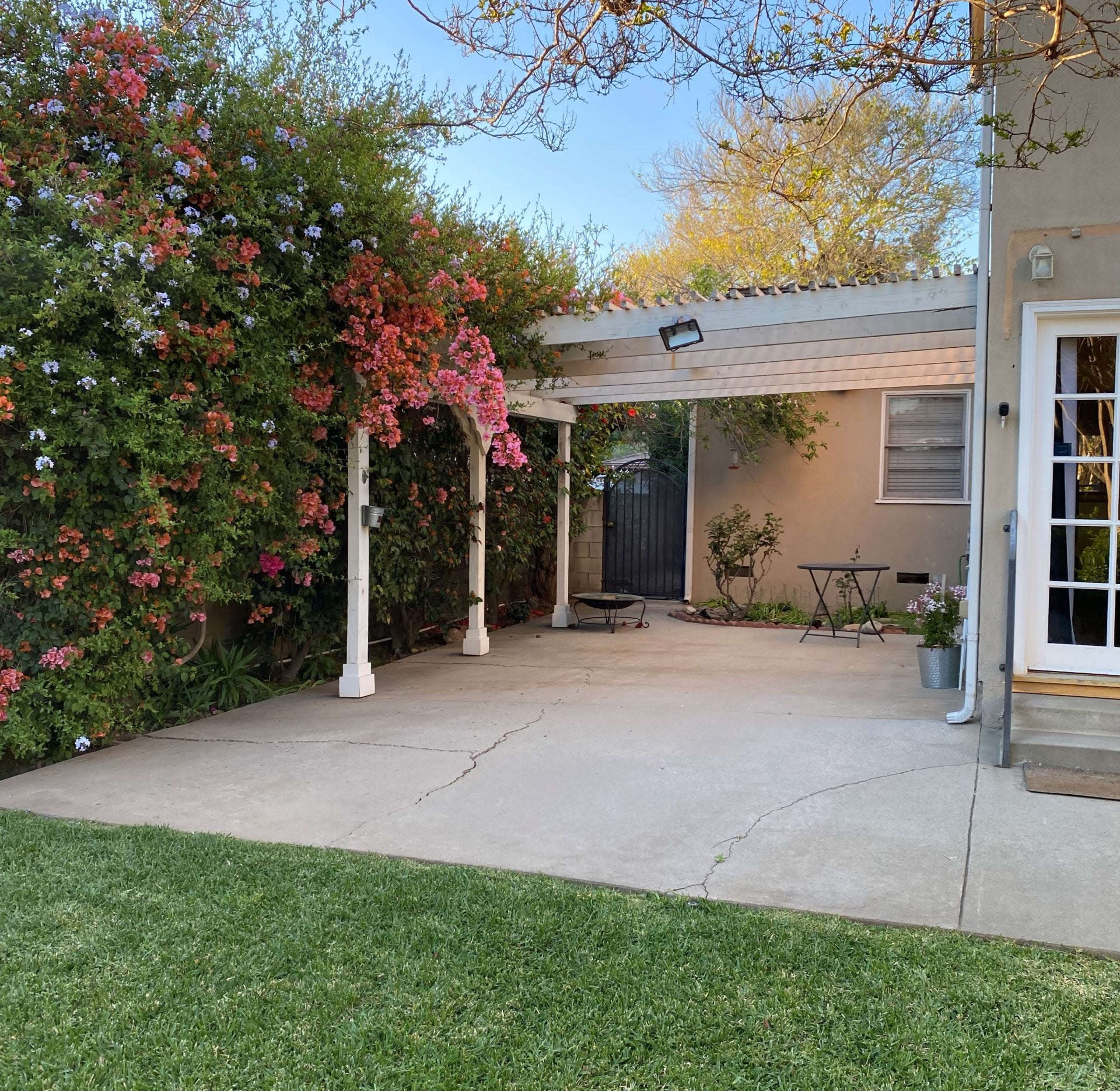 A patio area with a wooden trellis covered in flowering vines is situated next to a house, adjacent to a neatly trimmed lawn.