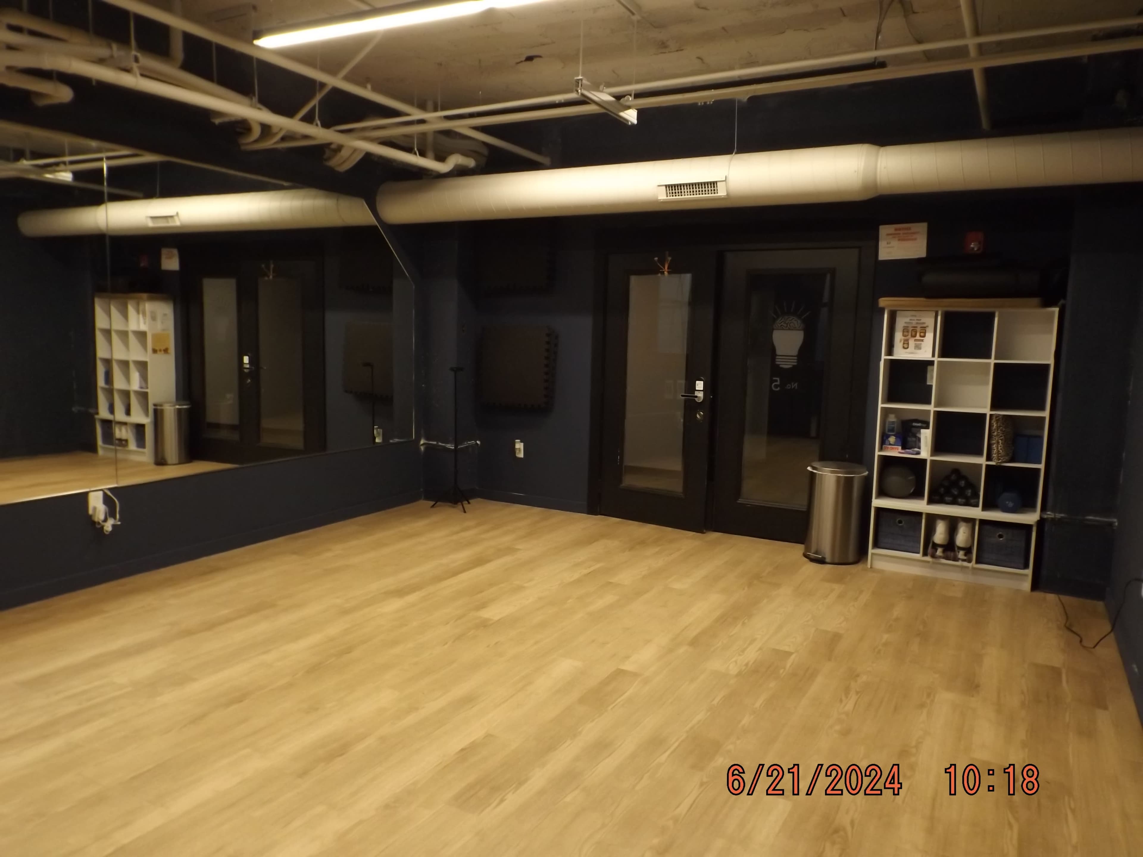 The image shows an empty room with wooden flooring, a large mirror on one wall, and black glass doors on the opposite side.