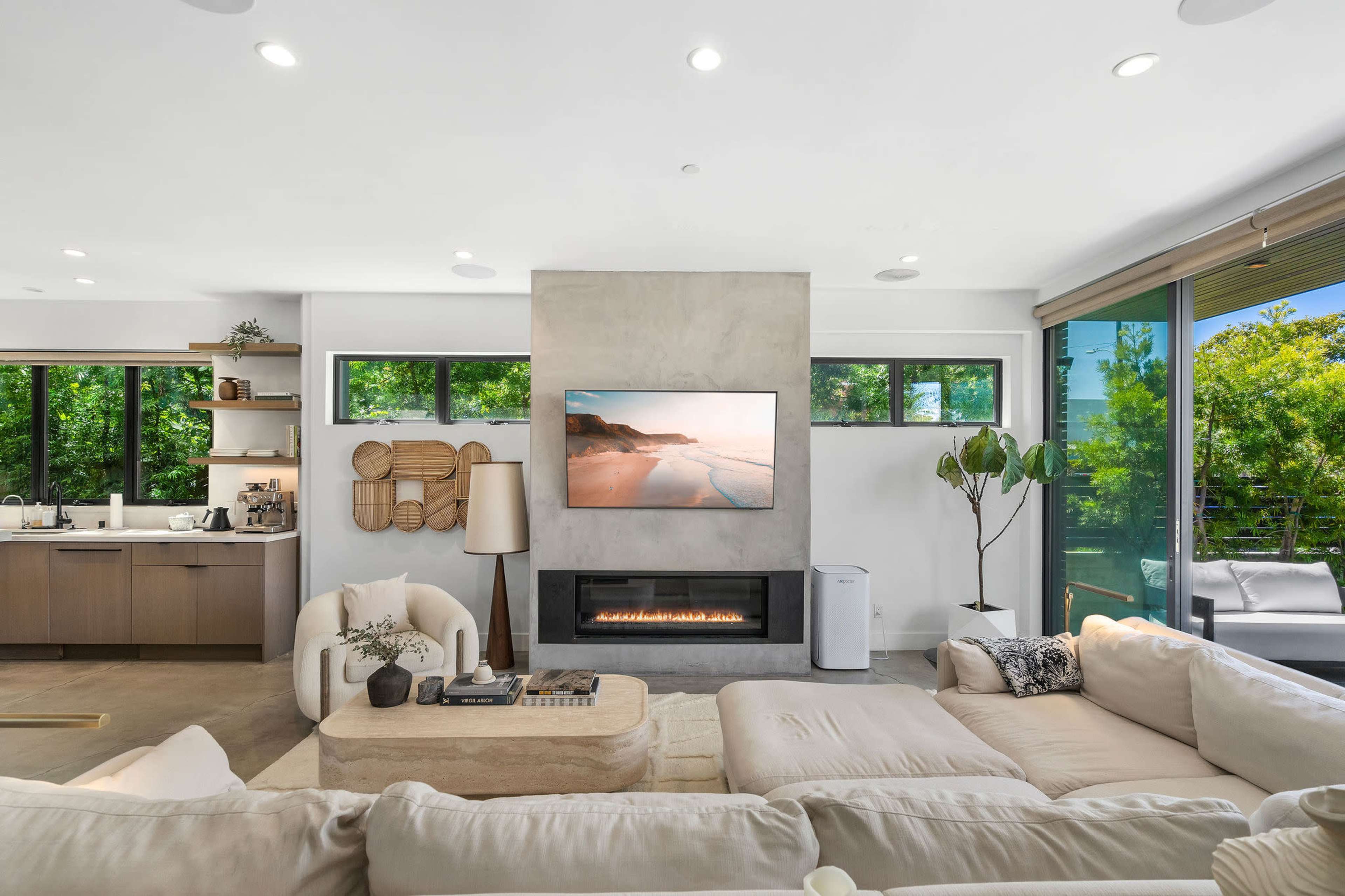 A modern living room with a large sectional sofa, a wall-mounted television above a fireplace, and windows that provide views of greenery outside.