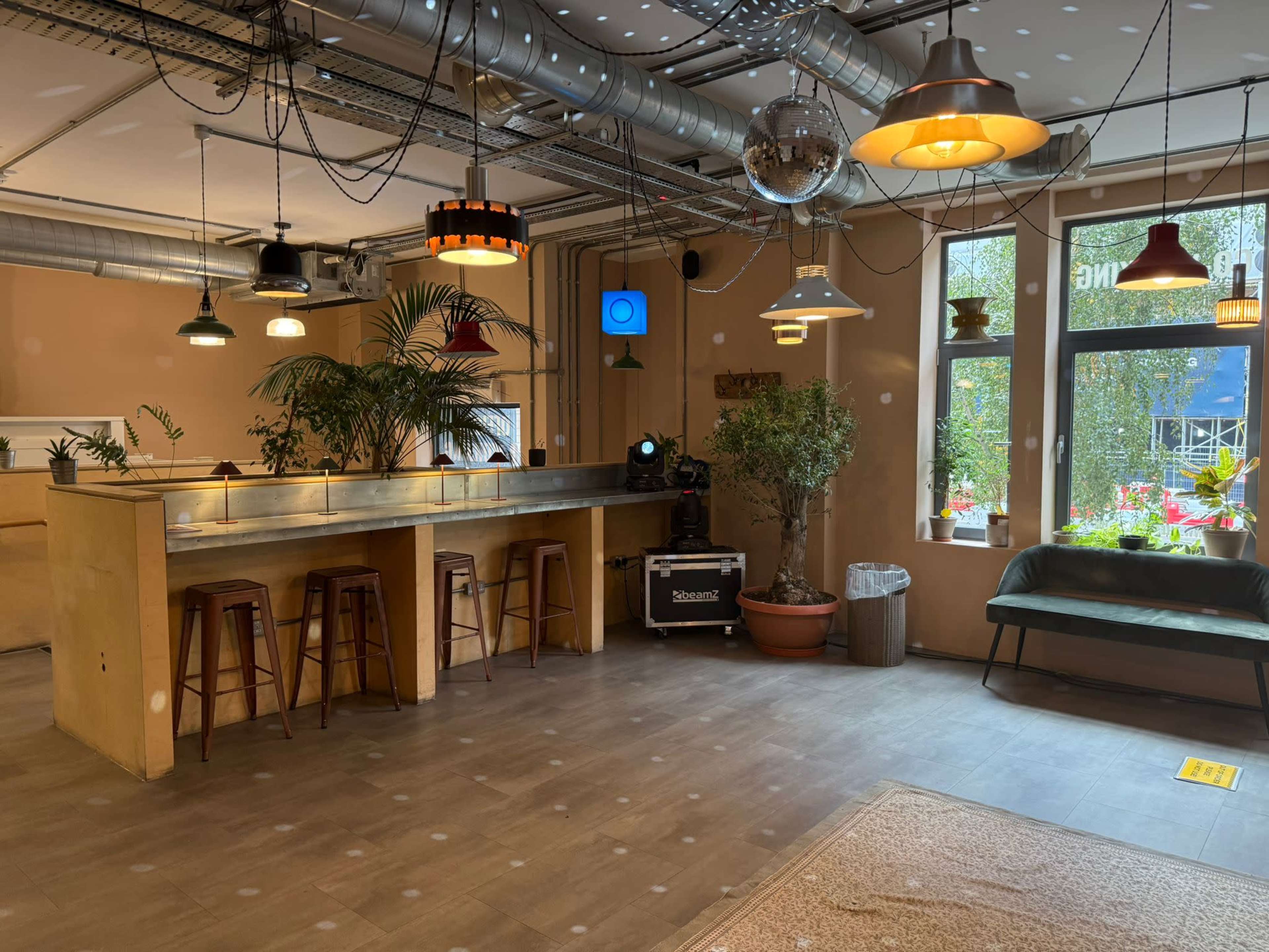 The image features a cozy interior space with a bar counter, a few bar stools, and plants, illuminated by various hanging lights.