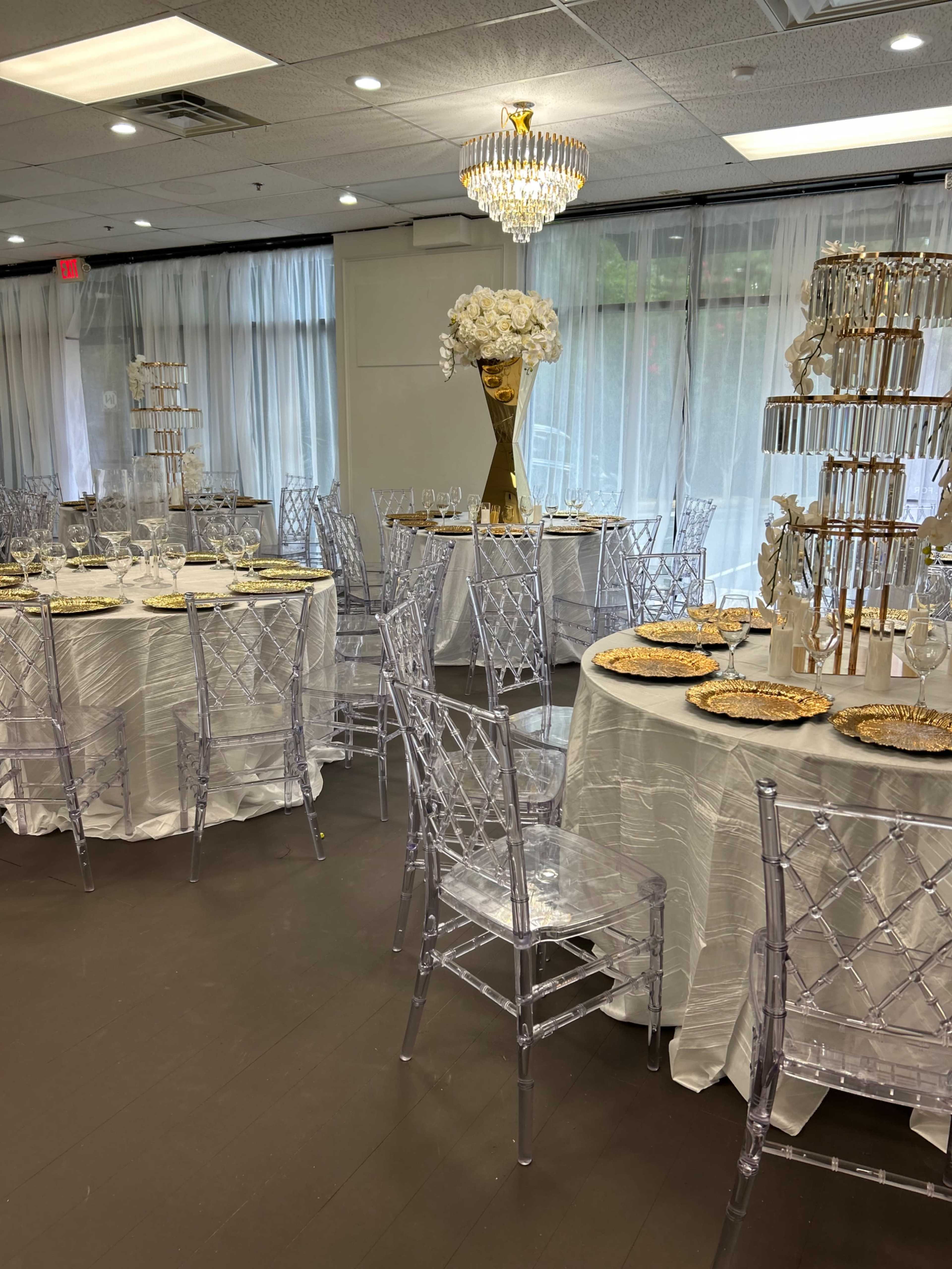 The image shows an elegantly arranged indoor event space with round tables dressed in white tablecloths, clear acrylic chairs, and floral centerpieces, illuminated by a chandelier.