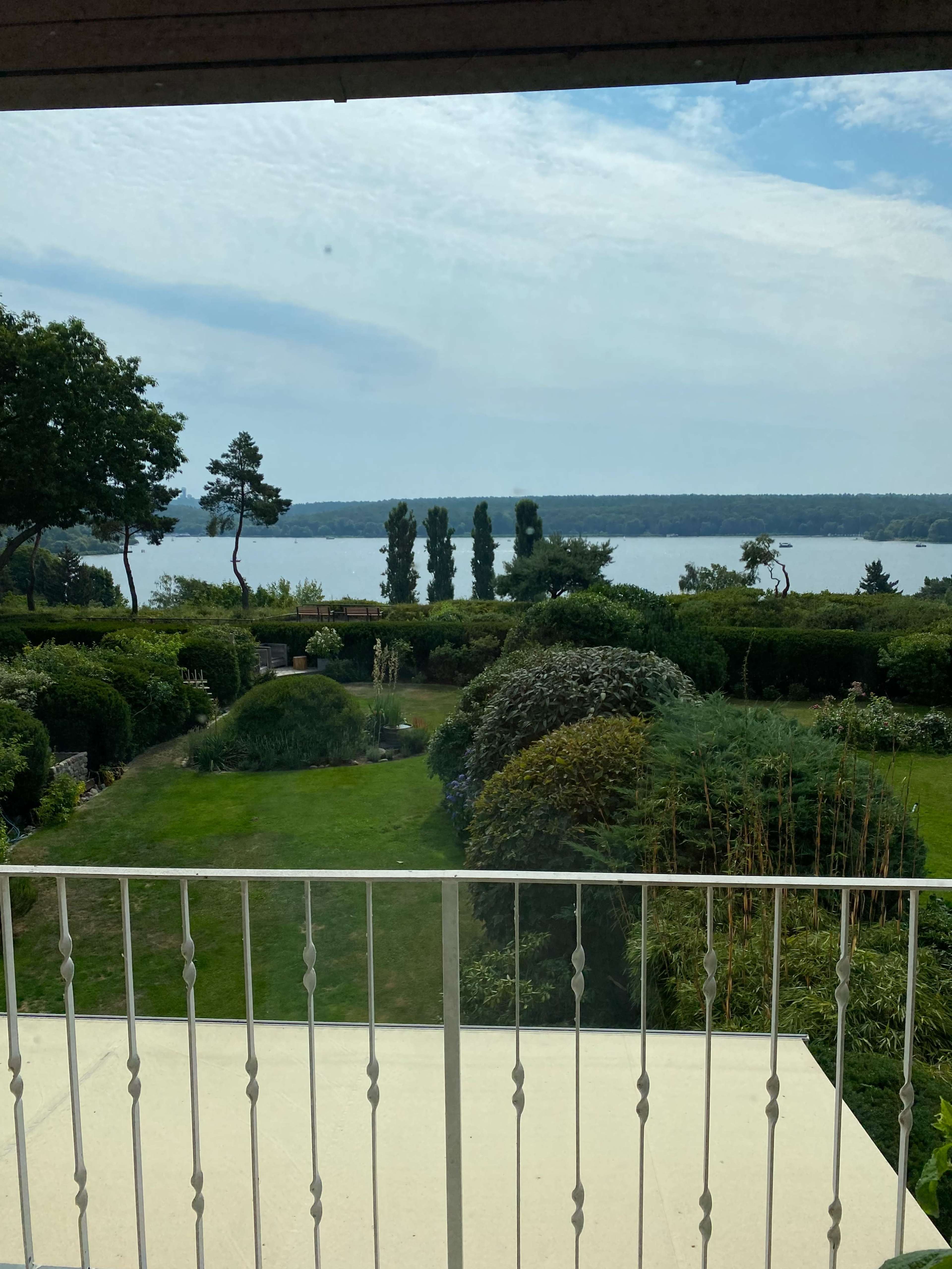 A view from a balcony overlooking a lush garden and a calm lake surrounded by trees.
