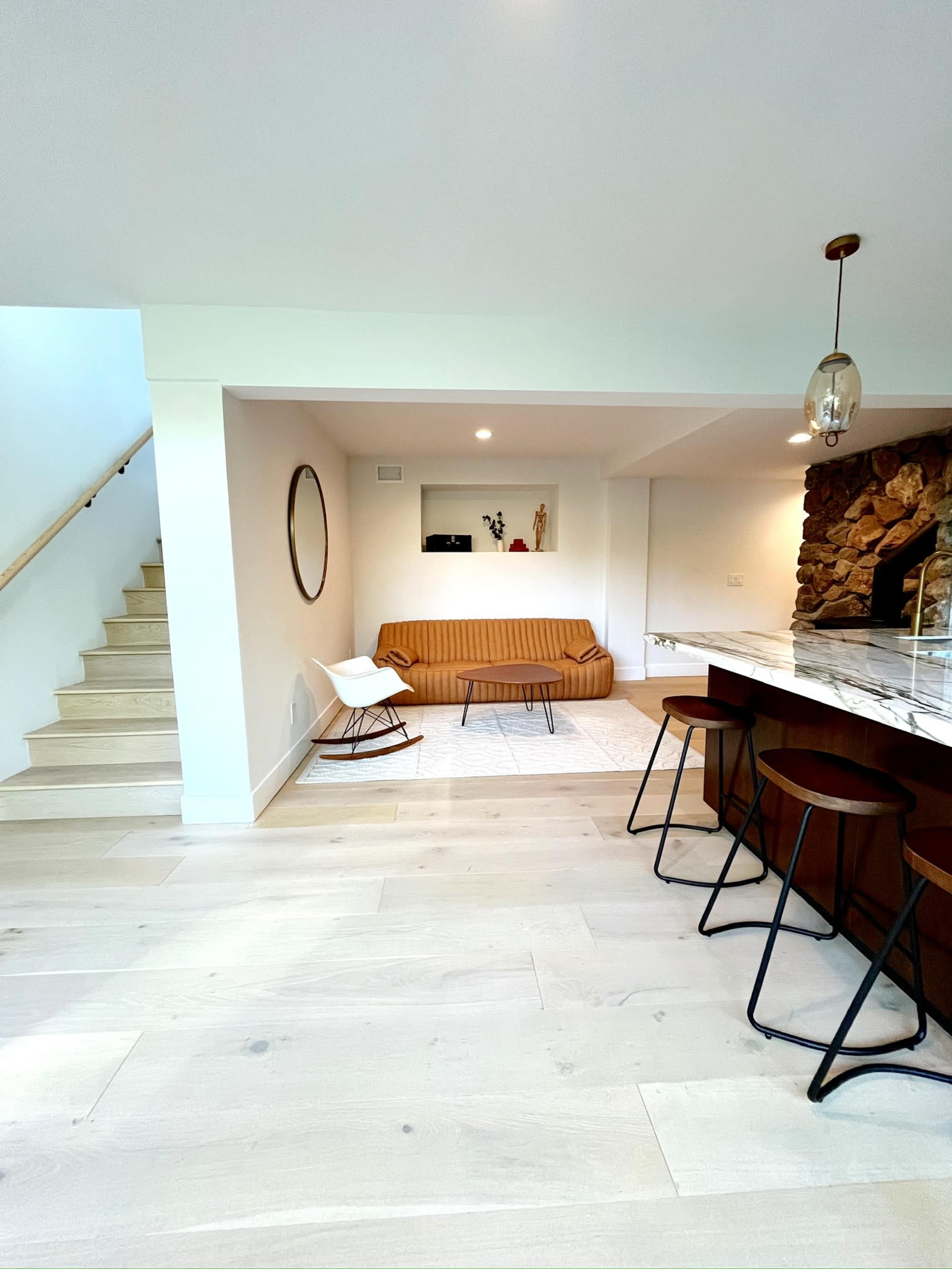 The image shows a modern living space with a sofa, a kitchen island with stools, a circular mirror on the wall, and a staircase in the background.