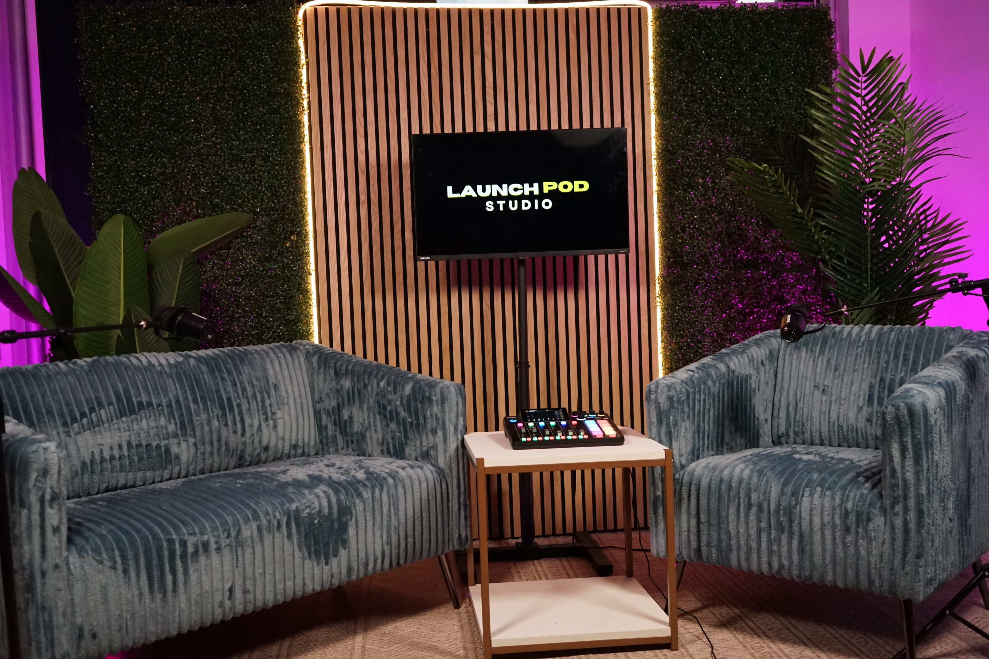 The image shows a modern podcasting setup with two blue velvet chairs facing a small white table, a screen displaying "LAUNCH POD STUDIO," and a backdrop of wooden slats and greenery.