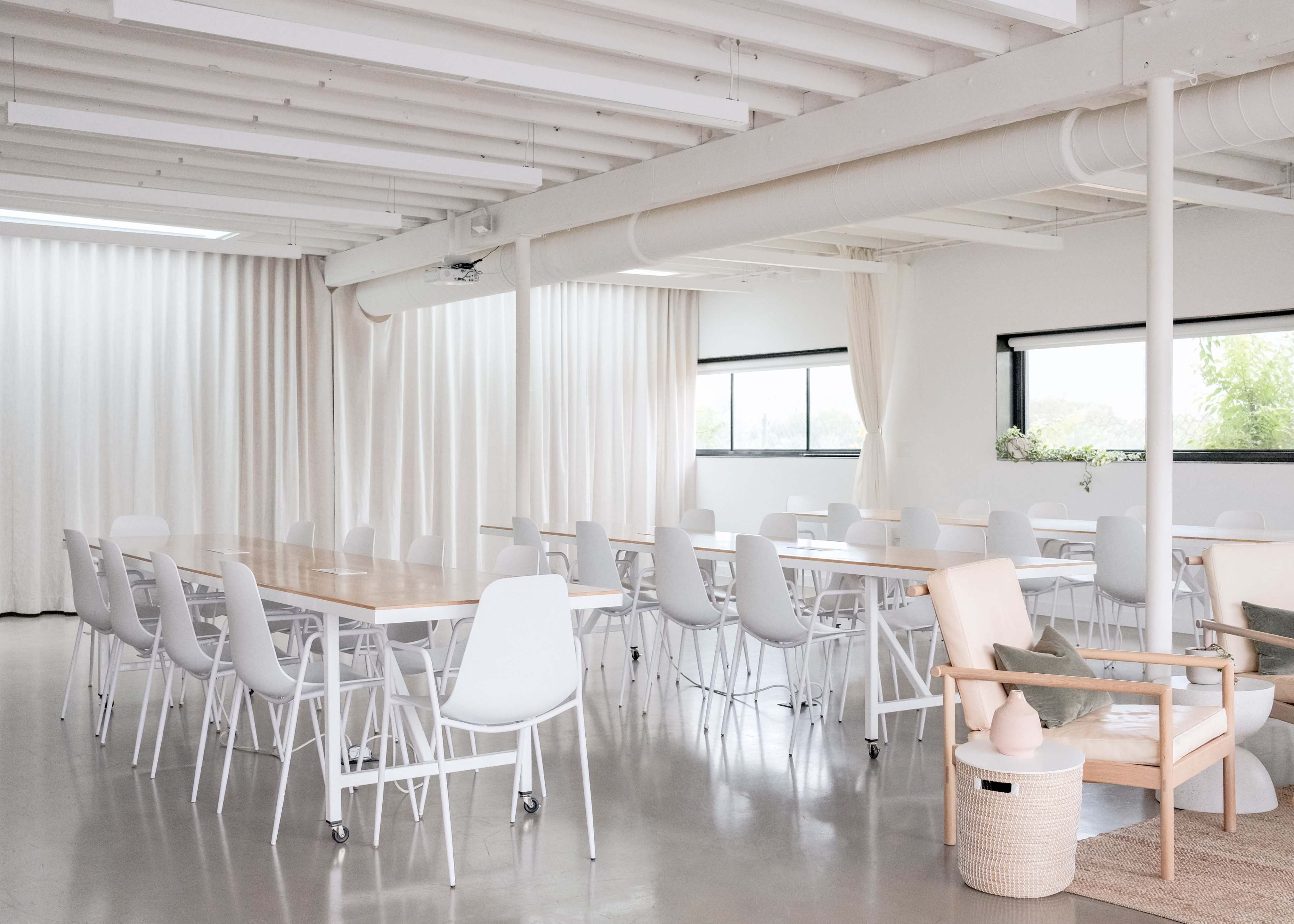 A spacious meeting room features long wooden tables surrounded by white chairs and large windows with sheer curtains.