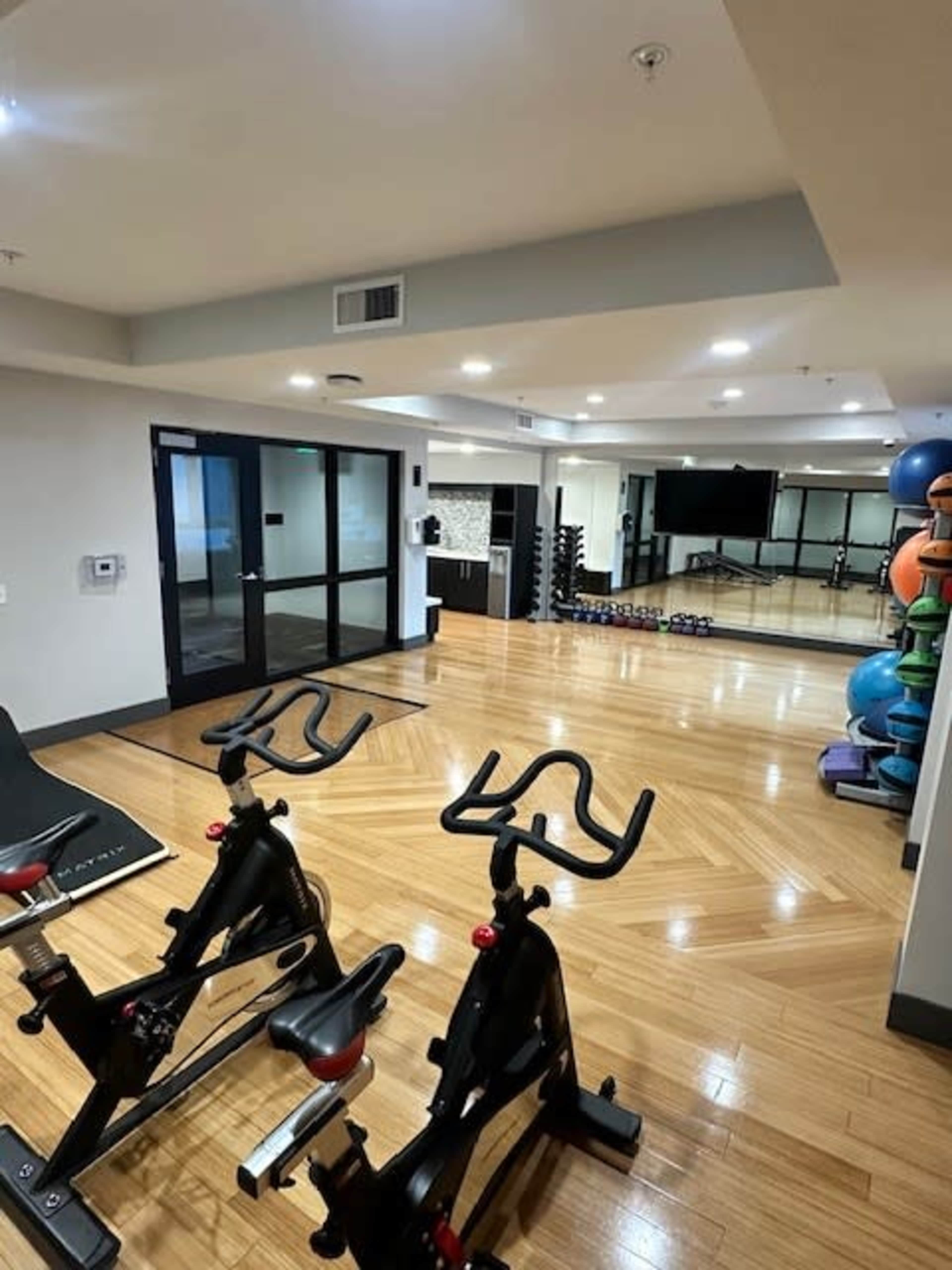 The image shows a fitness room equipped with exercise bikes, dumbbells, stability balls, and a large mirror, all set on a polished wooden floor.