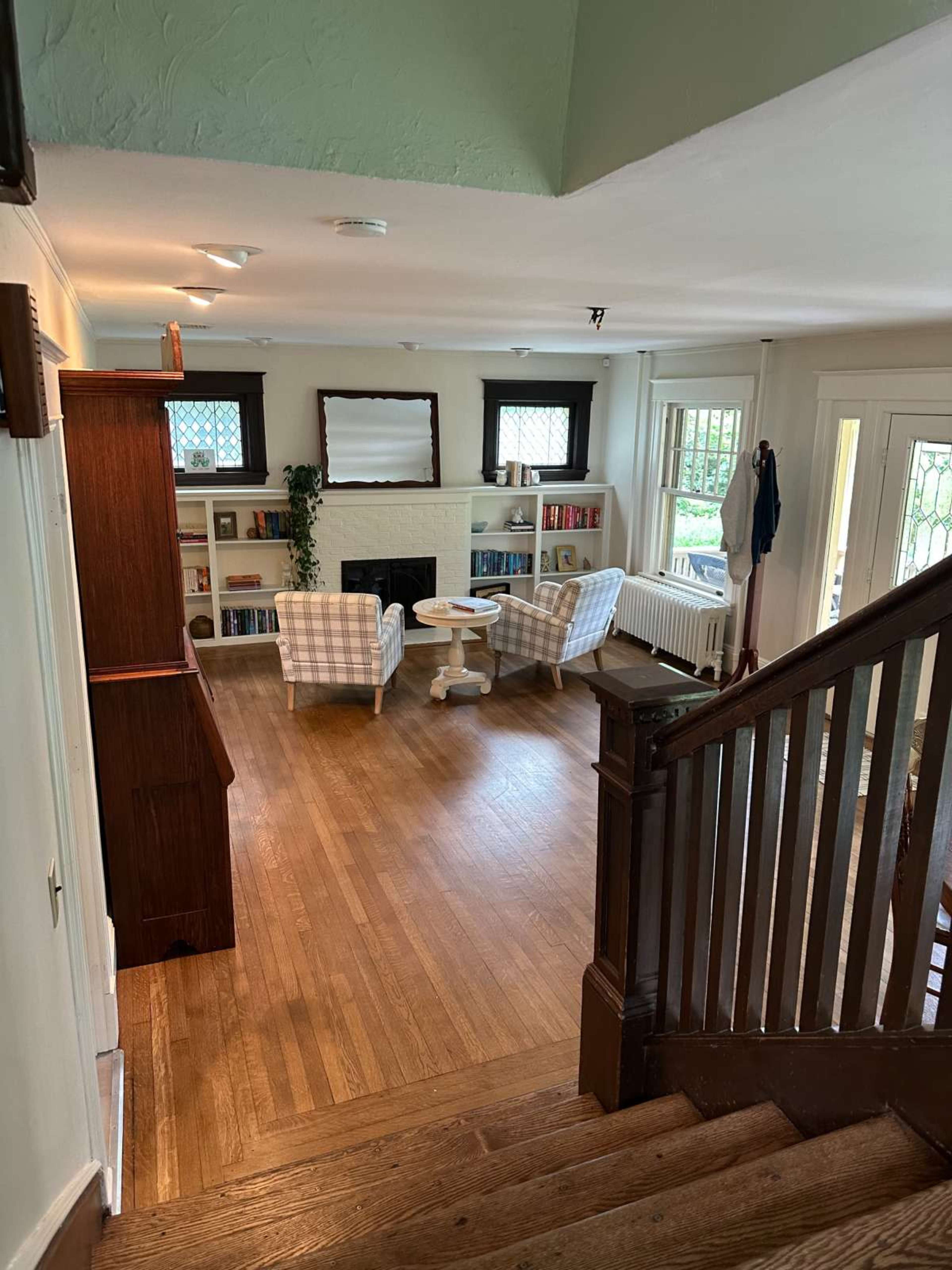 The image shows a well-lit living area with two armchairs facing a small table, a fireplace, and built-in bookshelves along the wall, as seen from a staircase.