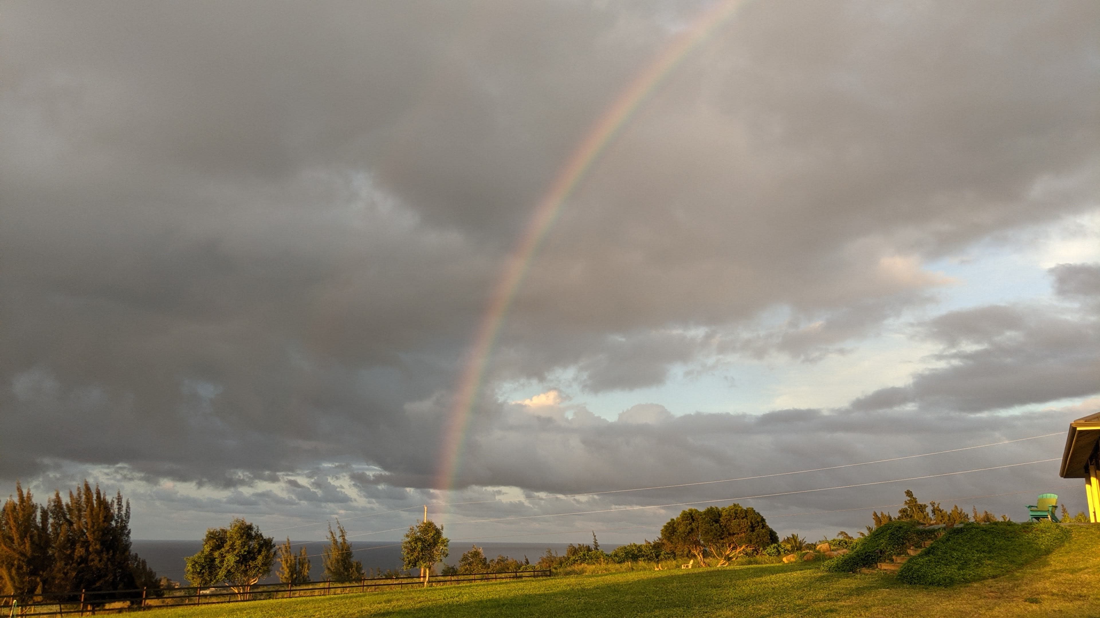 A rainbow arcs over a grassy field with dark clouds and a distant ocean in the background.