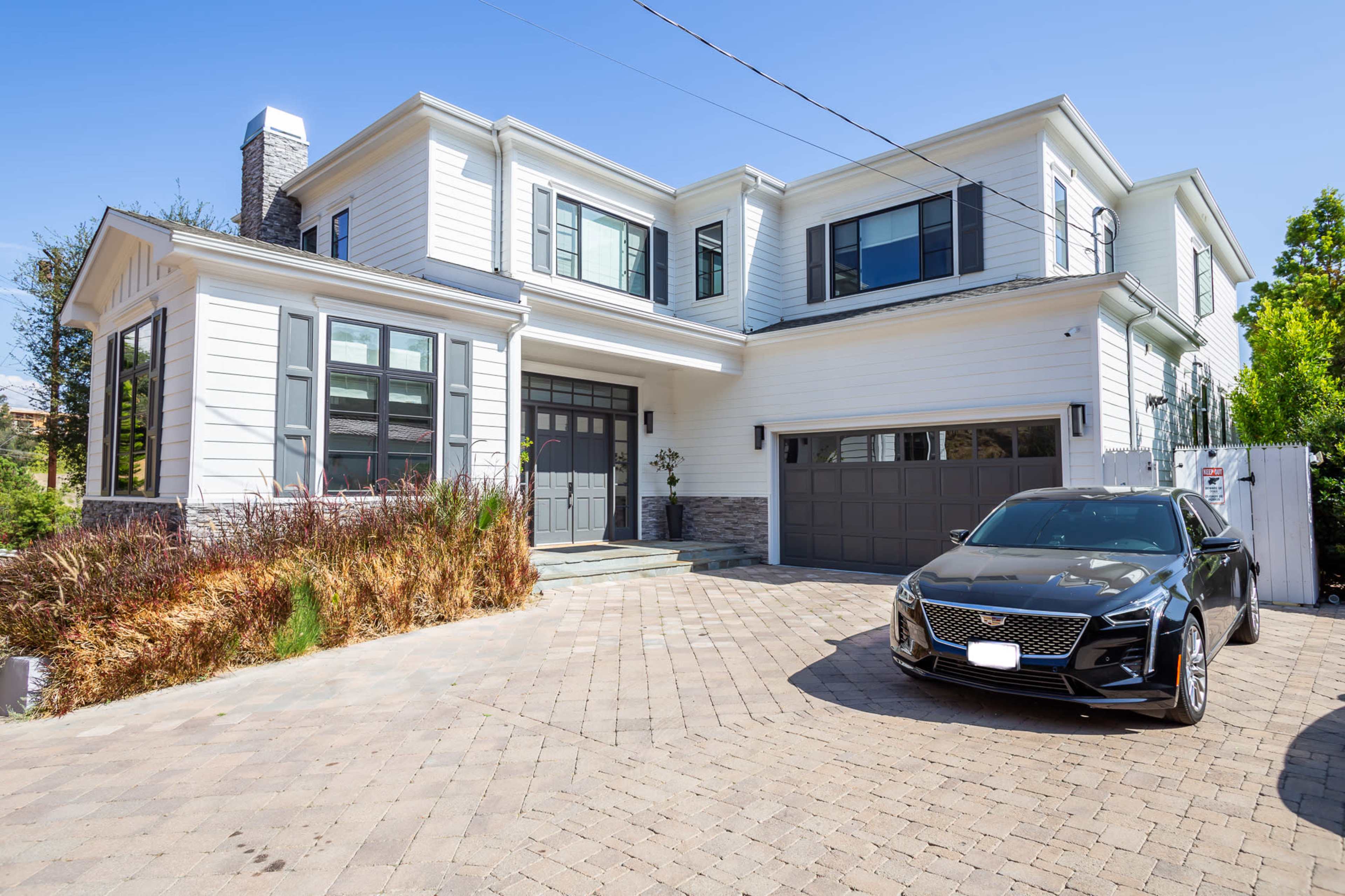 A modern two-story house with a gray and white exterior, large windows, and a driveway featuring a black car parked in front.