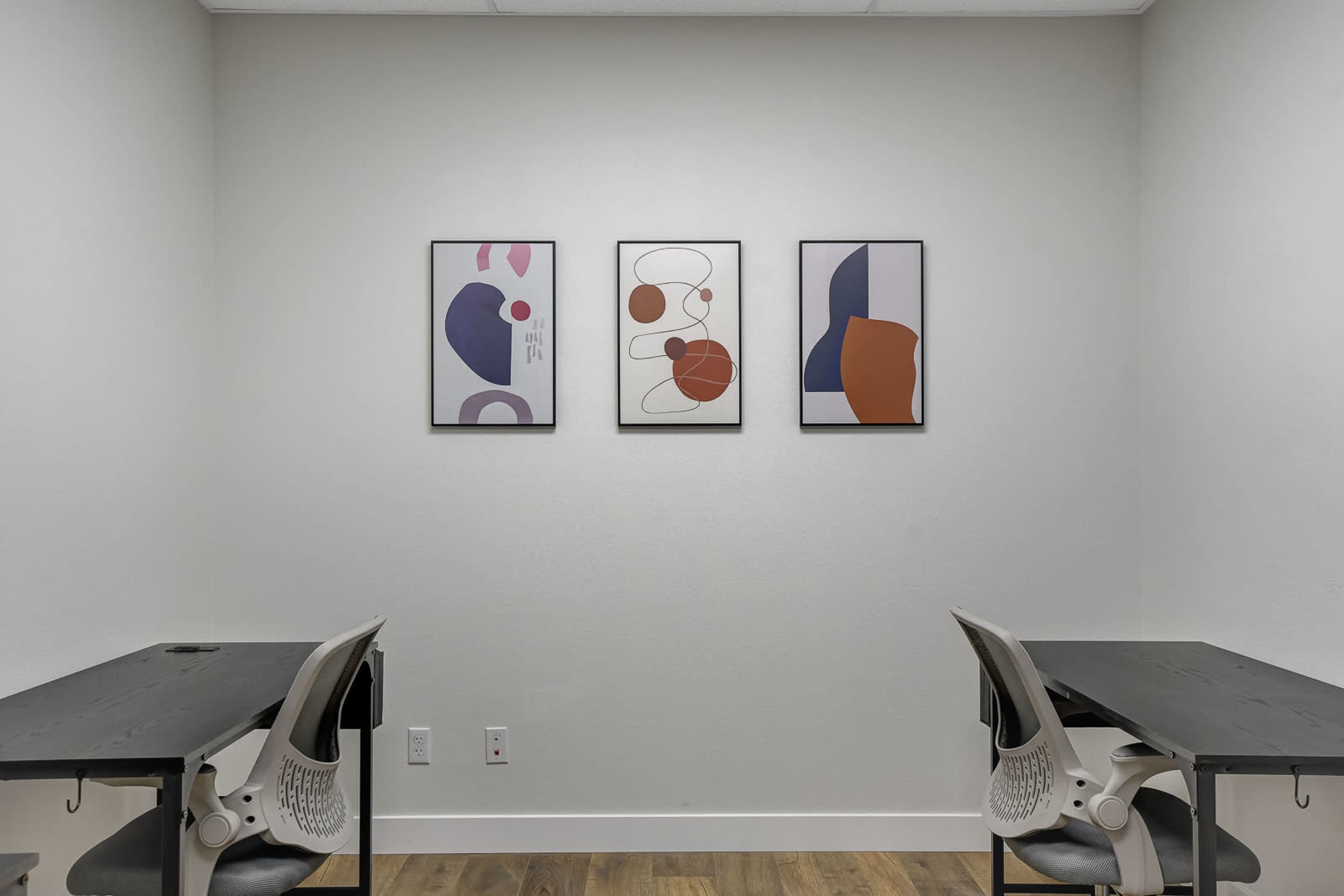 A minimalist office space with two black tables and three abstract art pieces hanging on a white wall.