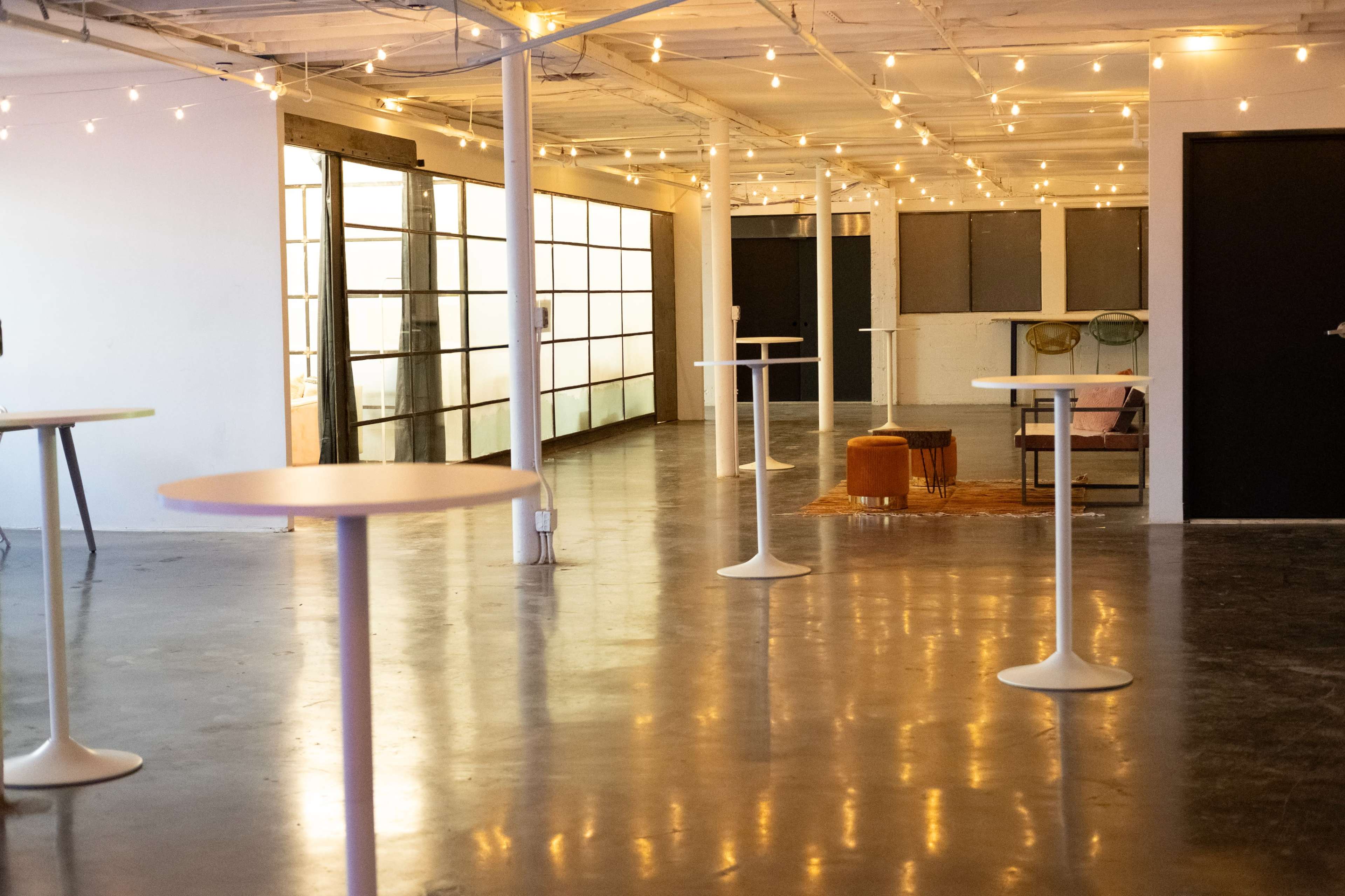 The image shows a spacious, well-lit interior with multiple tall, round tables and string lights illuminating the ceiling.