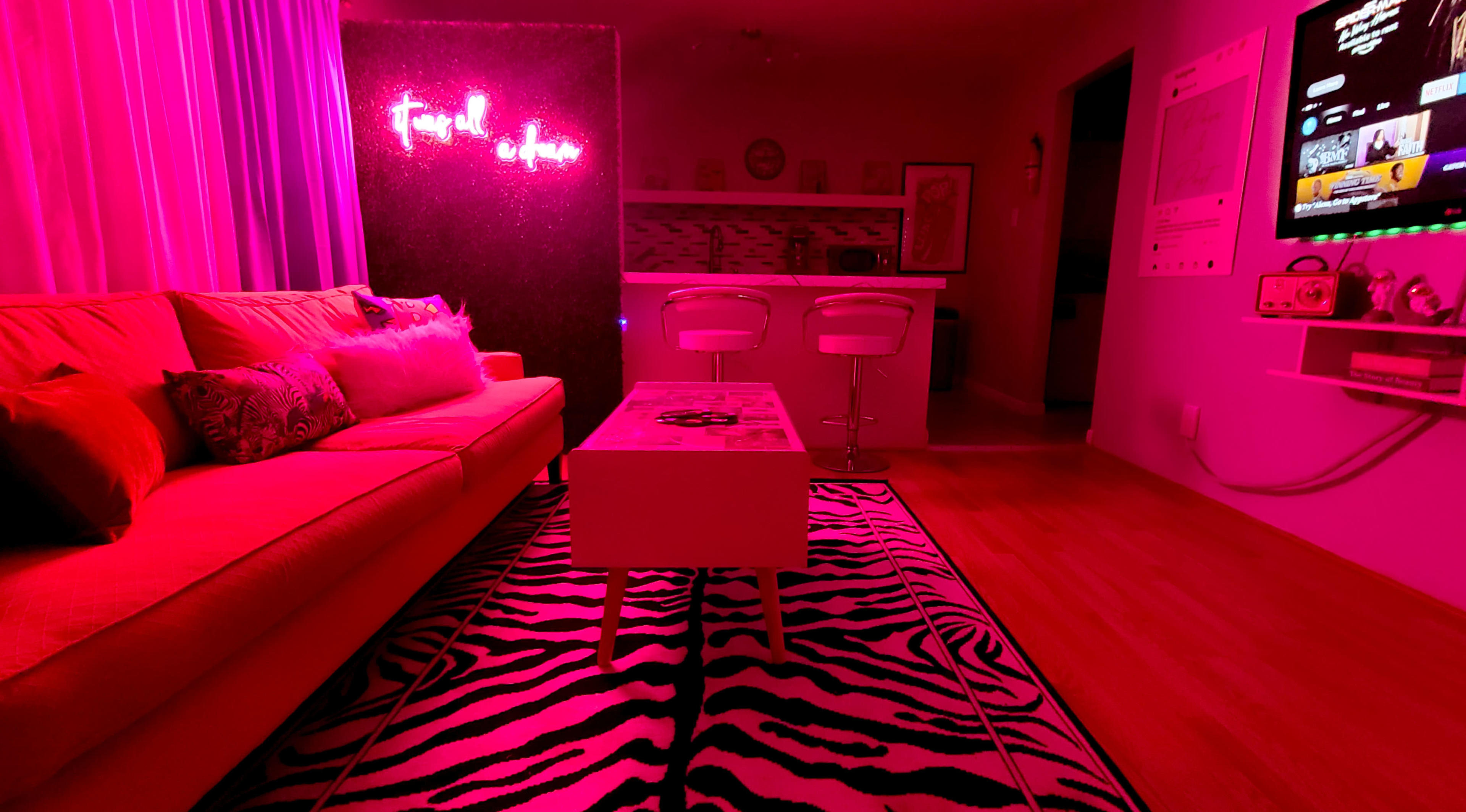 The image shows a brightly lit room with a red sofa, a zebra-striped rug, a modern coffee table, and a glowing neon sign on the wall.