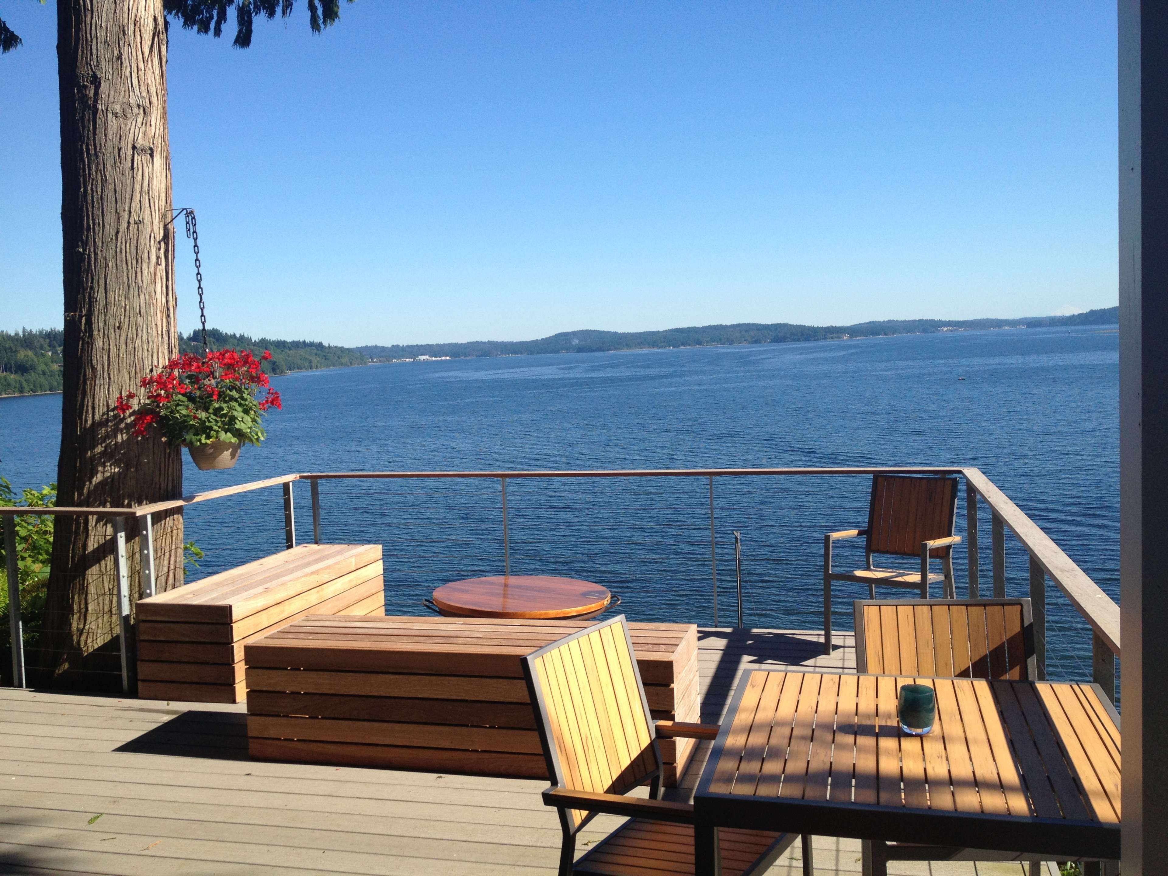 A deck overlooking a calm lake is furnished with wooden seating and a small table, surrounded by trees and distant hills.