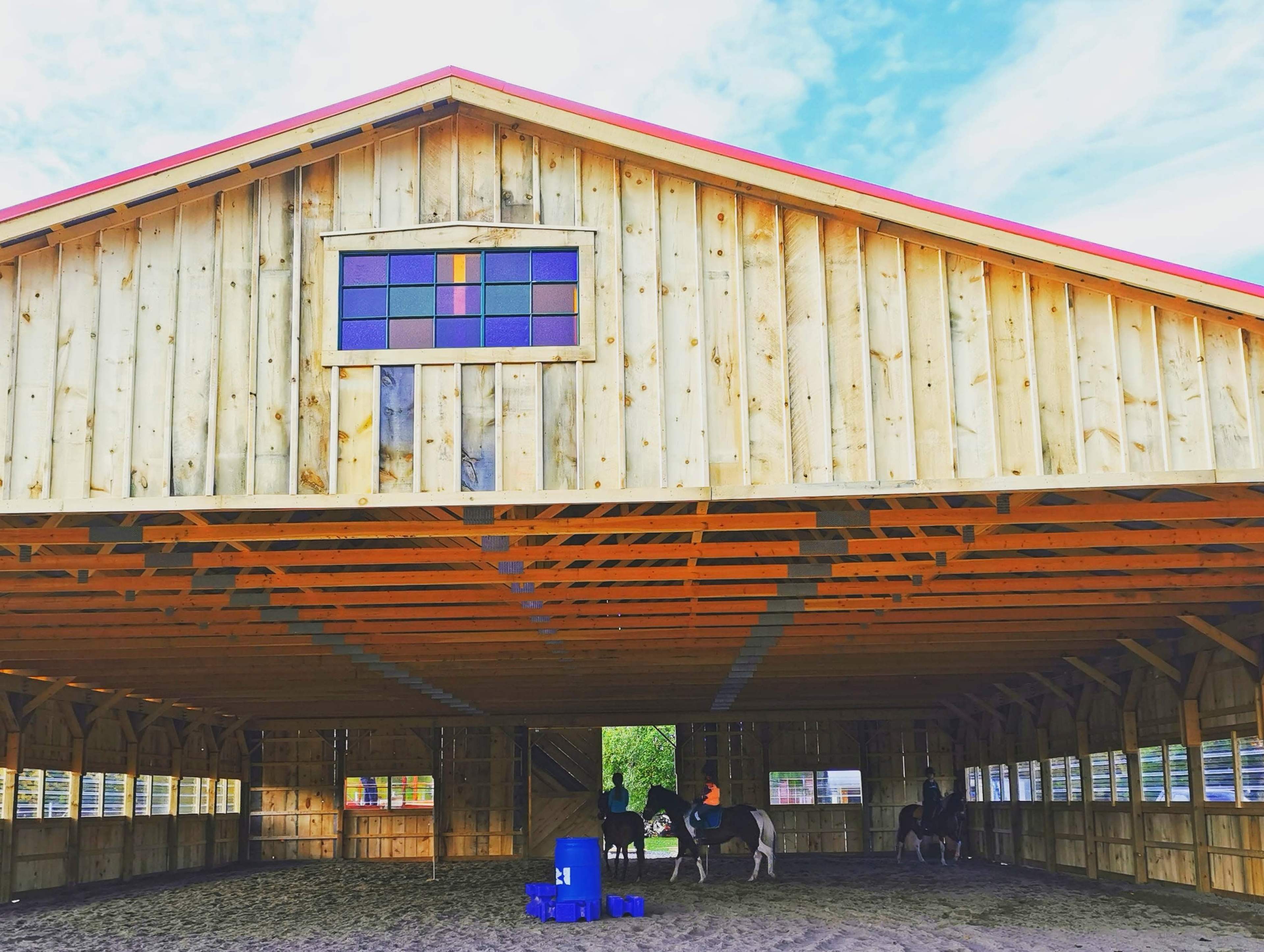 The image shows a wooden horse barn with a colorful stained glass window, where two horses are being ridden inside.