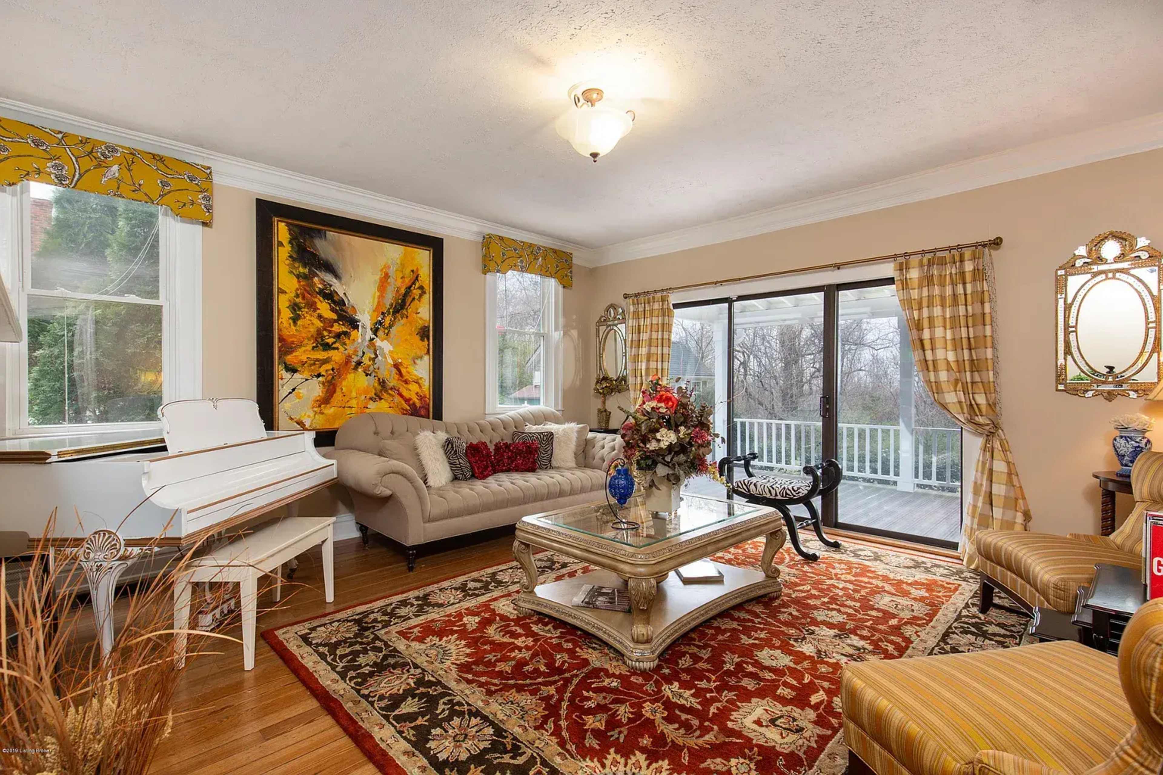 The room features a beige color scheme with a piano, a decorative coffee table, a large area rug, and large windows overlooking a porch.