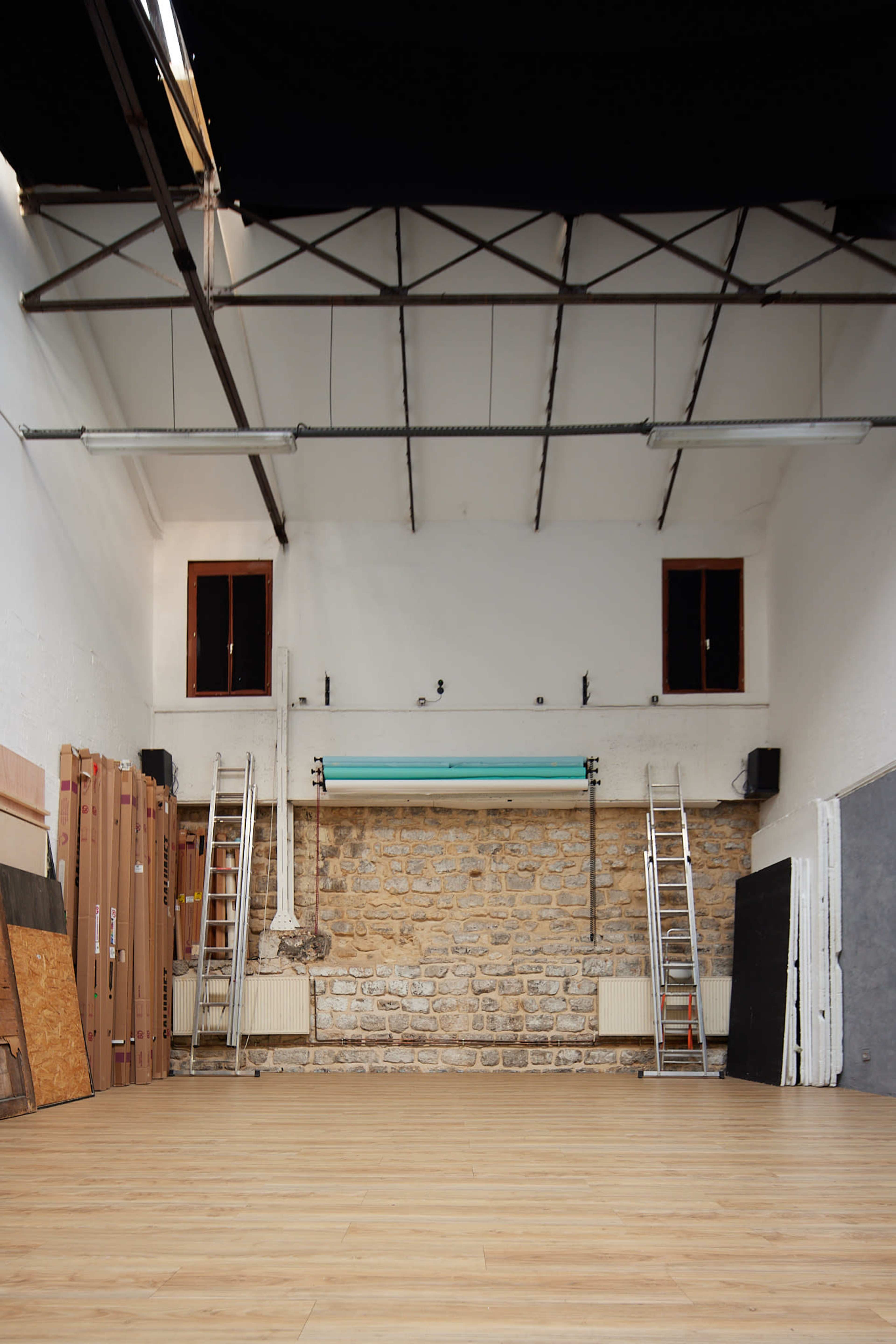 An empty room with wooden flooring, exposed stone walls, and ladders leaning against them, under a high ceiling with large windows.
