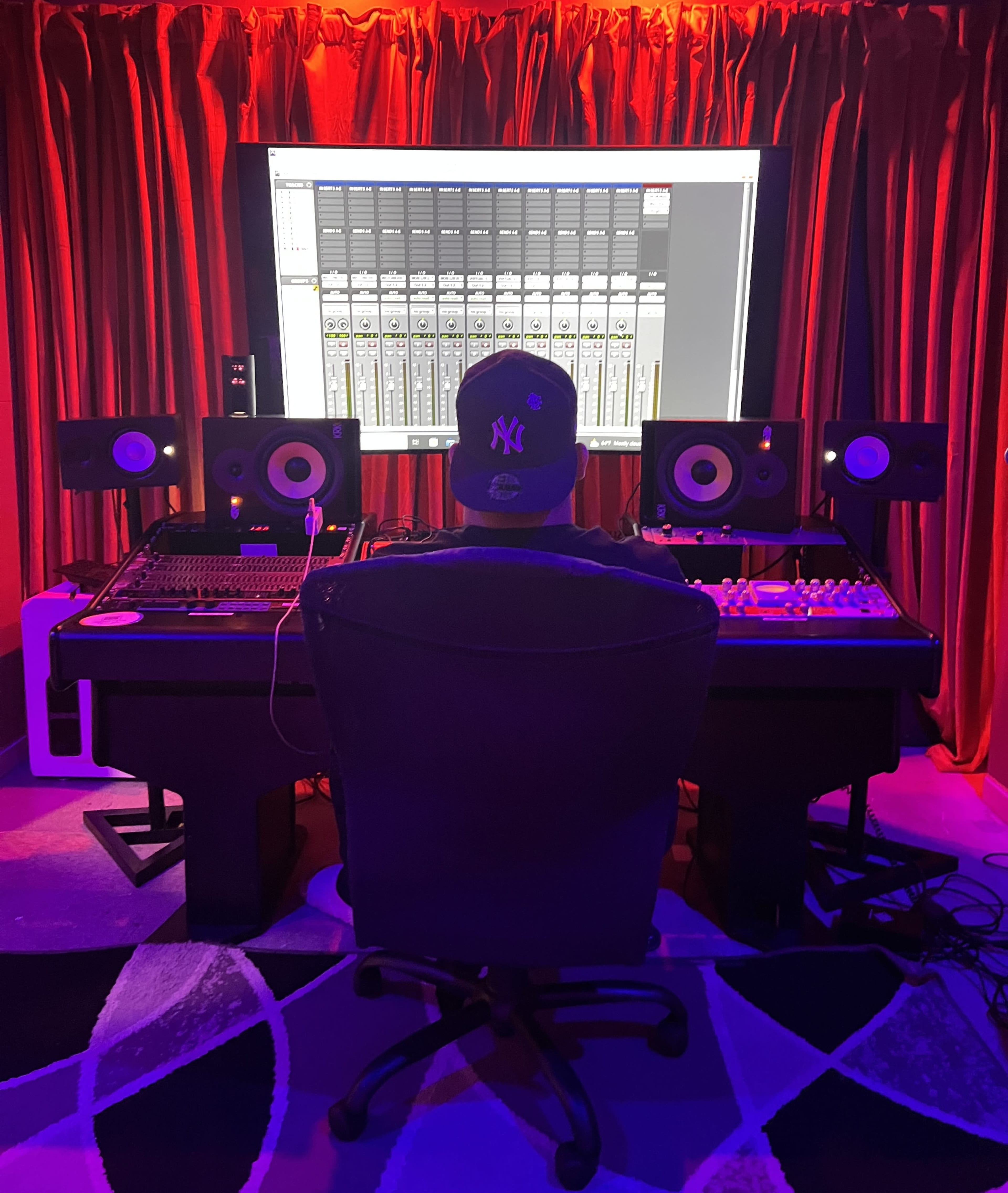 A person sits in a chair facing a large computer screen displaying audio mixing software in a recording studio.