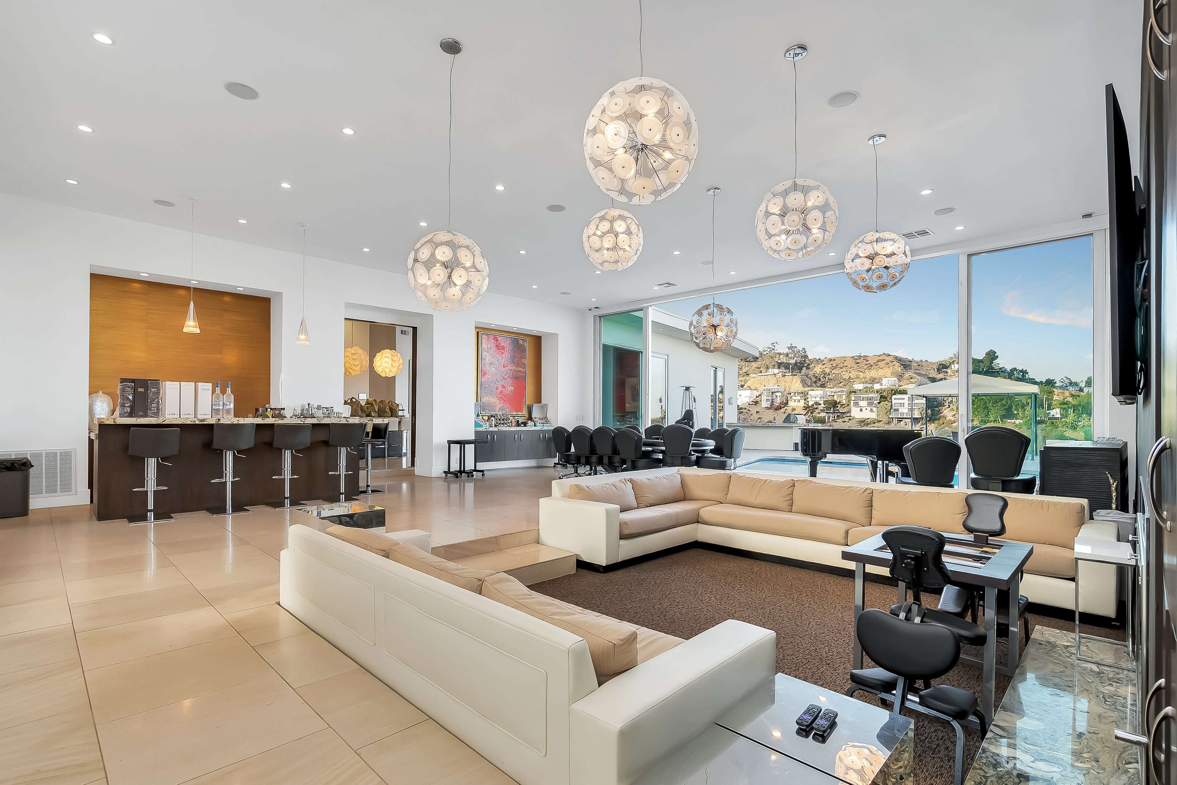 The image shows a spacious modern living area featuring a large sectional sofa, a bar area, and multiple decorative light fixtures, with large windows offering a view of the outdoors.