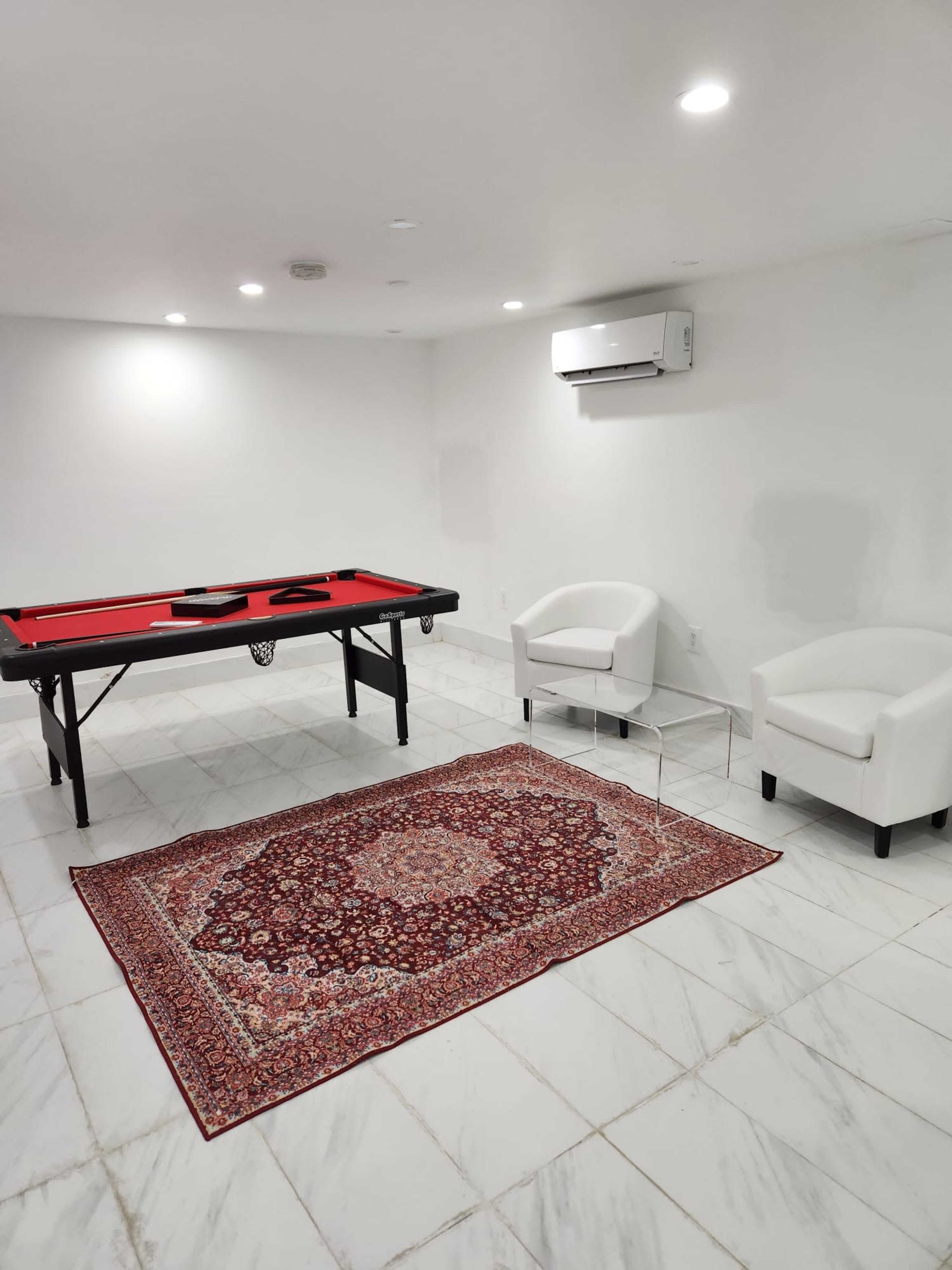 The image shows a room with a red pool table, two white chairs, and a patterned rug on a marble floor.