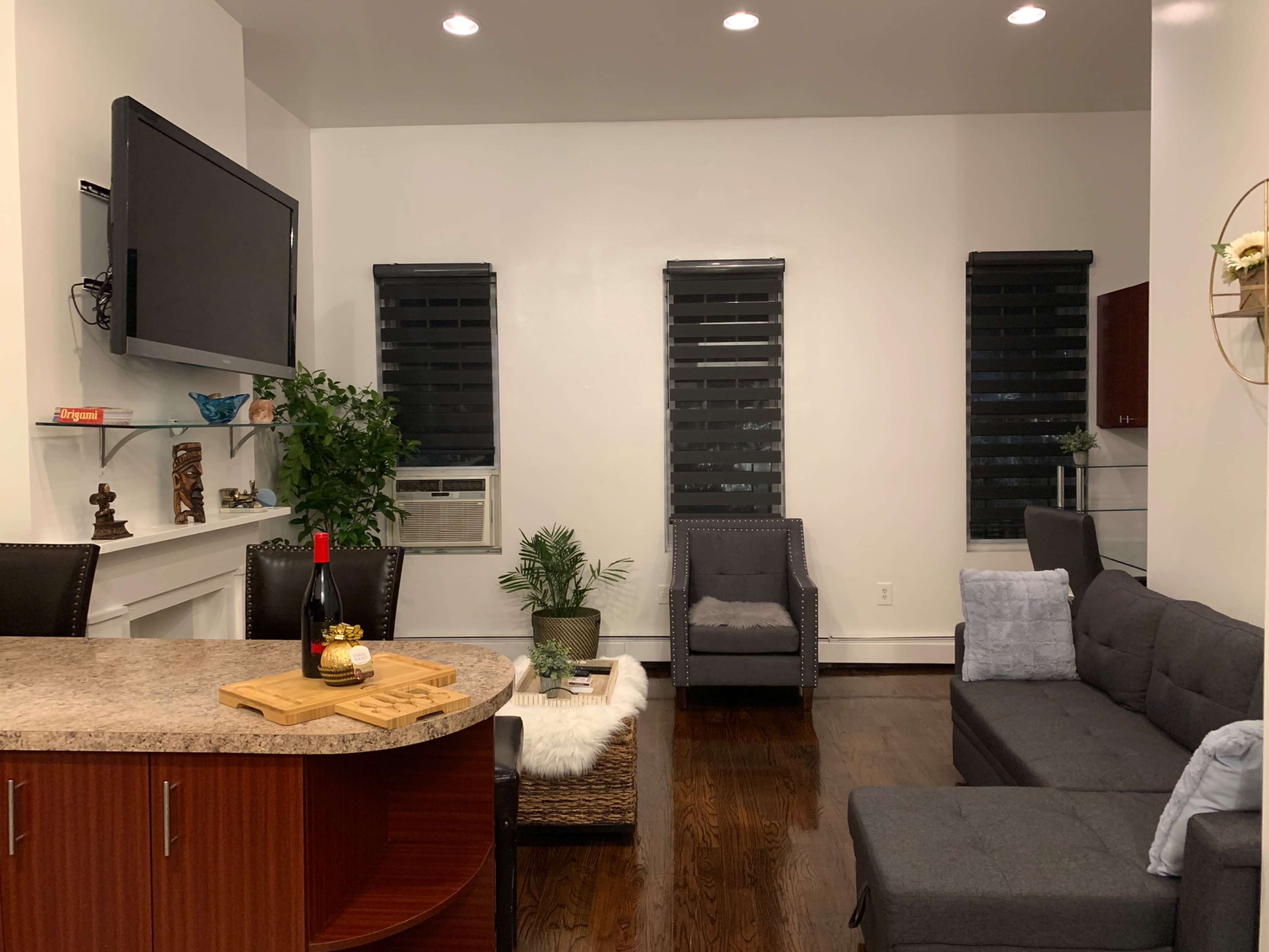 A modern living area with a dark gray sofa, two armchairs, a wall-mounted television, and a small kitchen counter with a bottle of wine and decorative elements.
