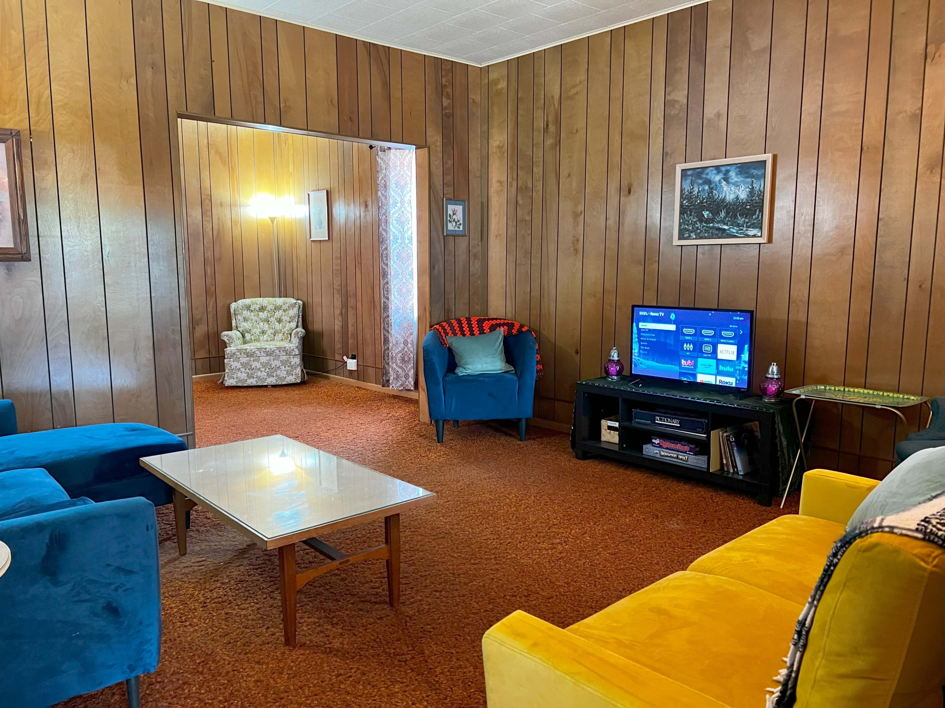 The image shows a living room with wooden paneling, a television on a black stand, a glass coffee table, and two colorful chairs across from a patterned armchair in an adjacent space.