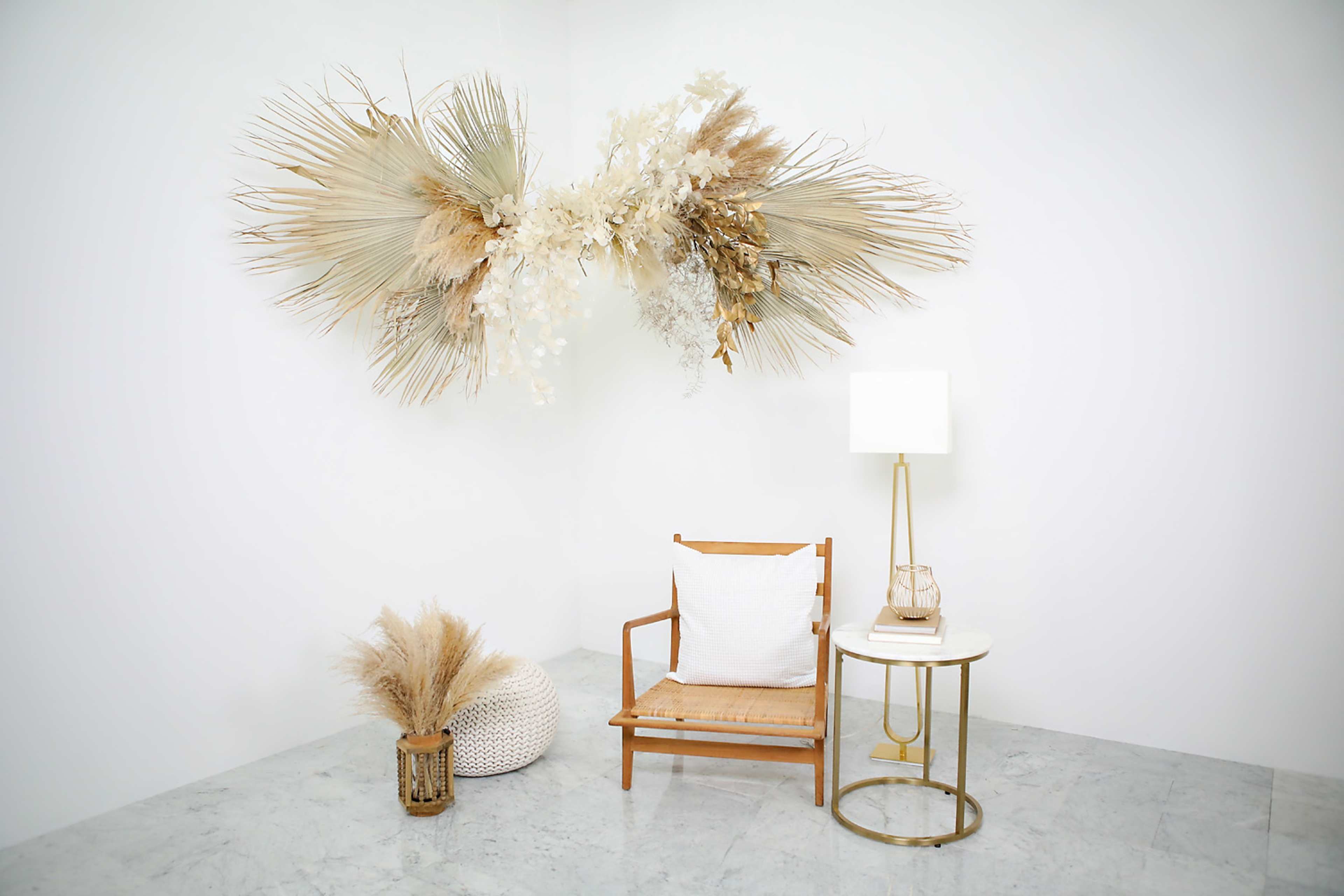 A minimalist interior design features a wooden chair with a cushion, a small white side table with a lamp, and decorative dried floral arrangements on the wall and floor.