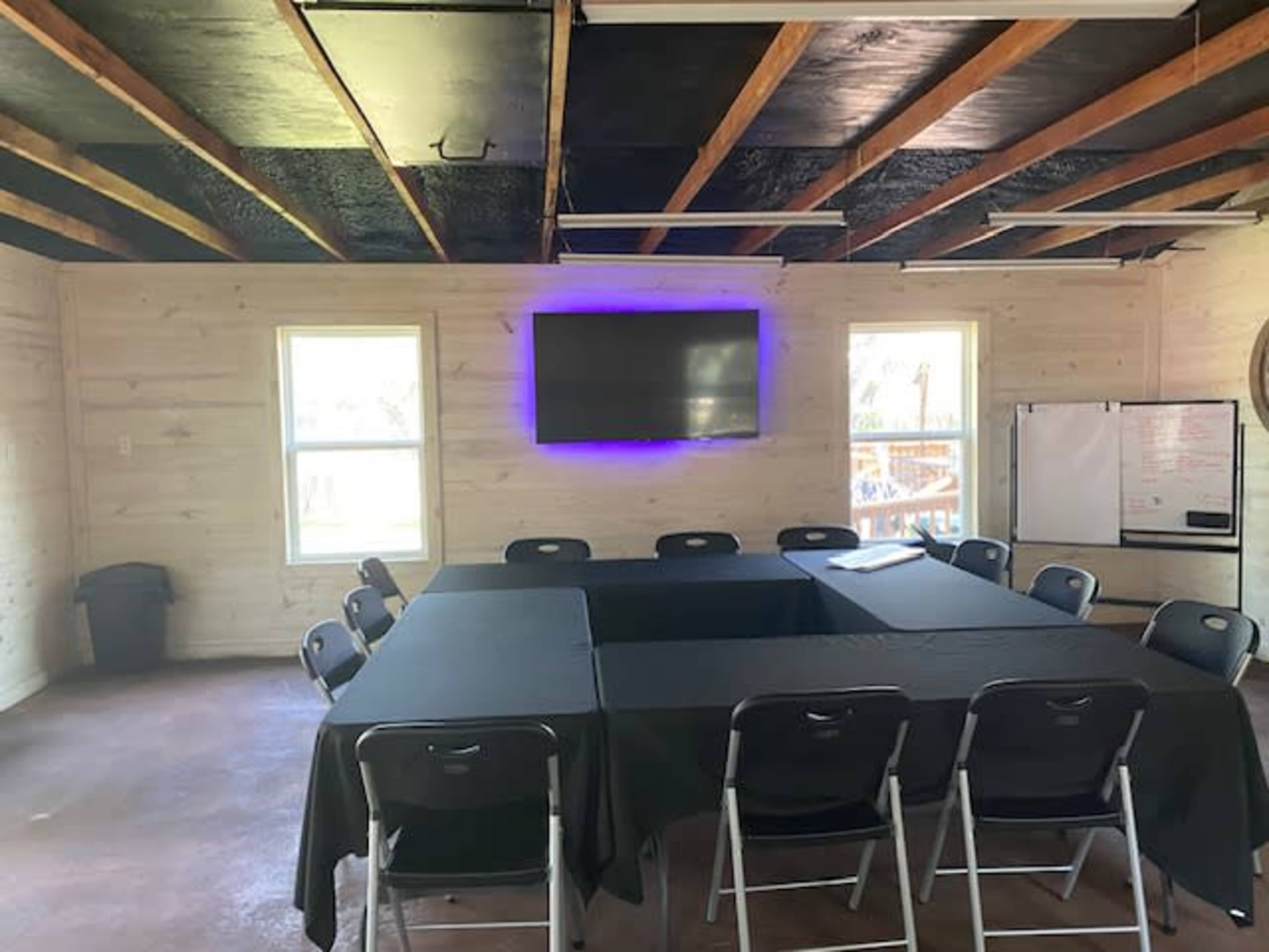 A conference room with a large table surrounded by chairs, featuring a television on the wall and windows allowing natural light.