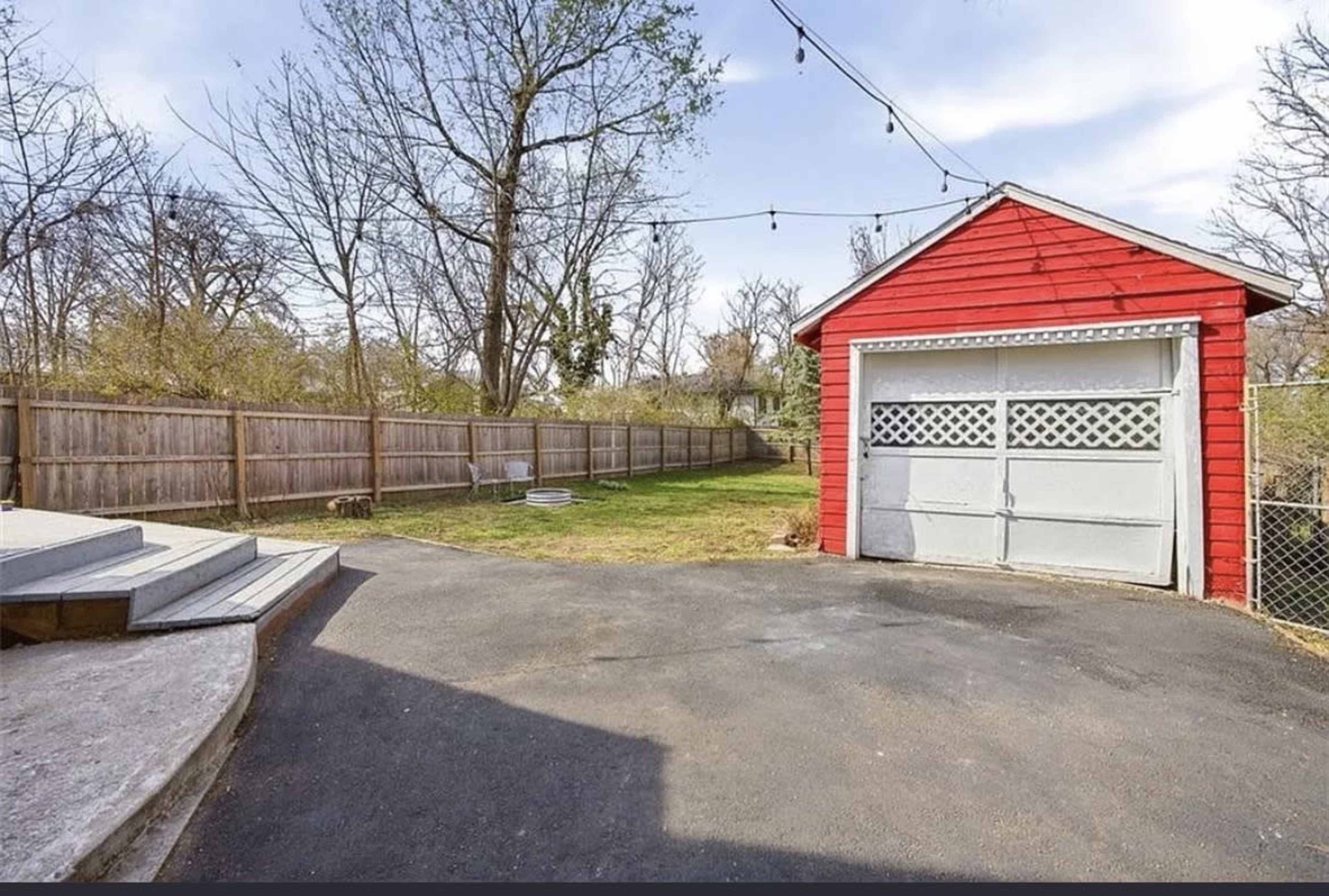The image shows a red garage beside a paved area in a yard surrounded by a wooden fence and sparse trees.