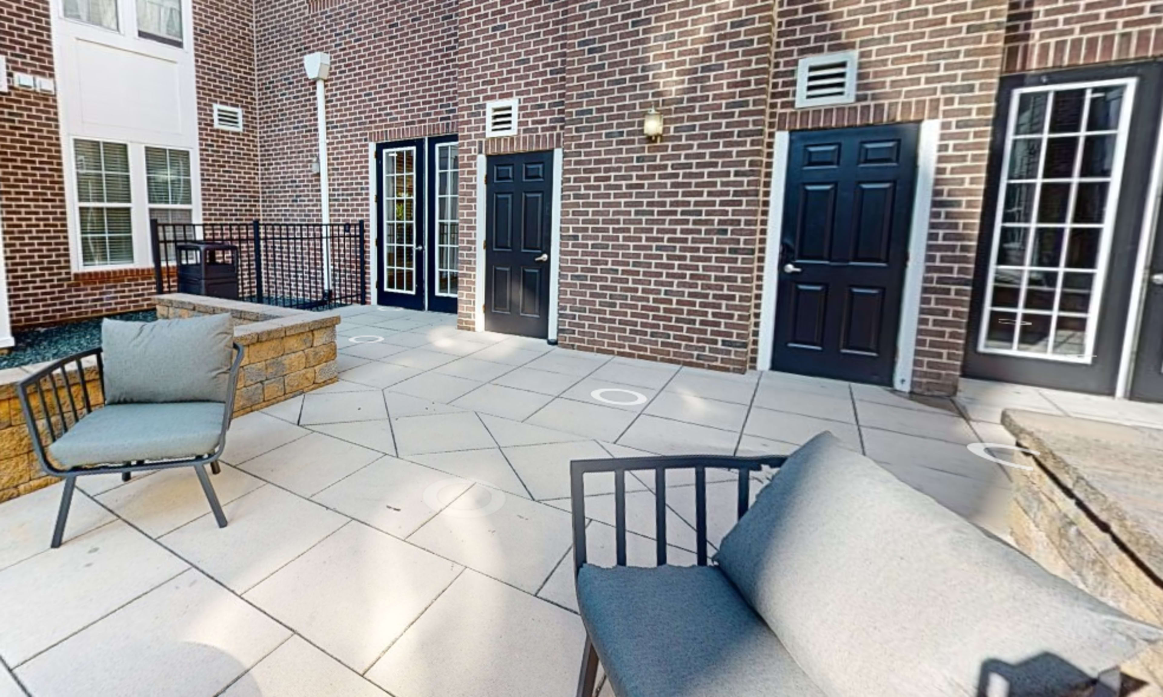 The image shows a patio area with two gray chairs, a stone wall, and doors leading into a building, all set against a brick wall.