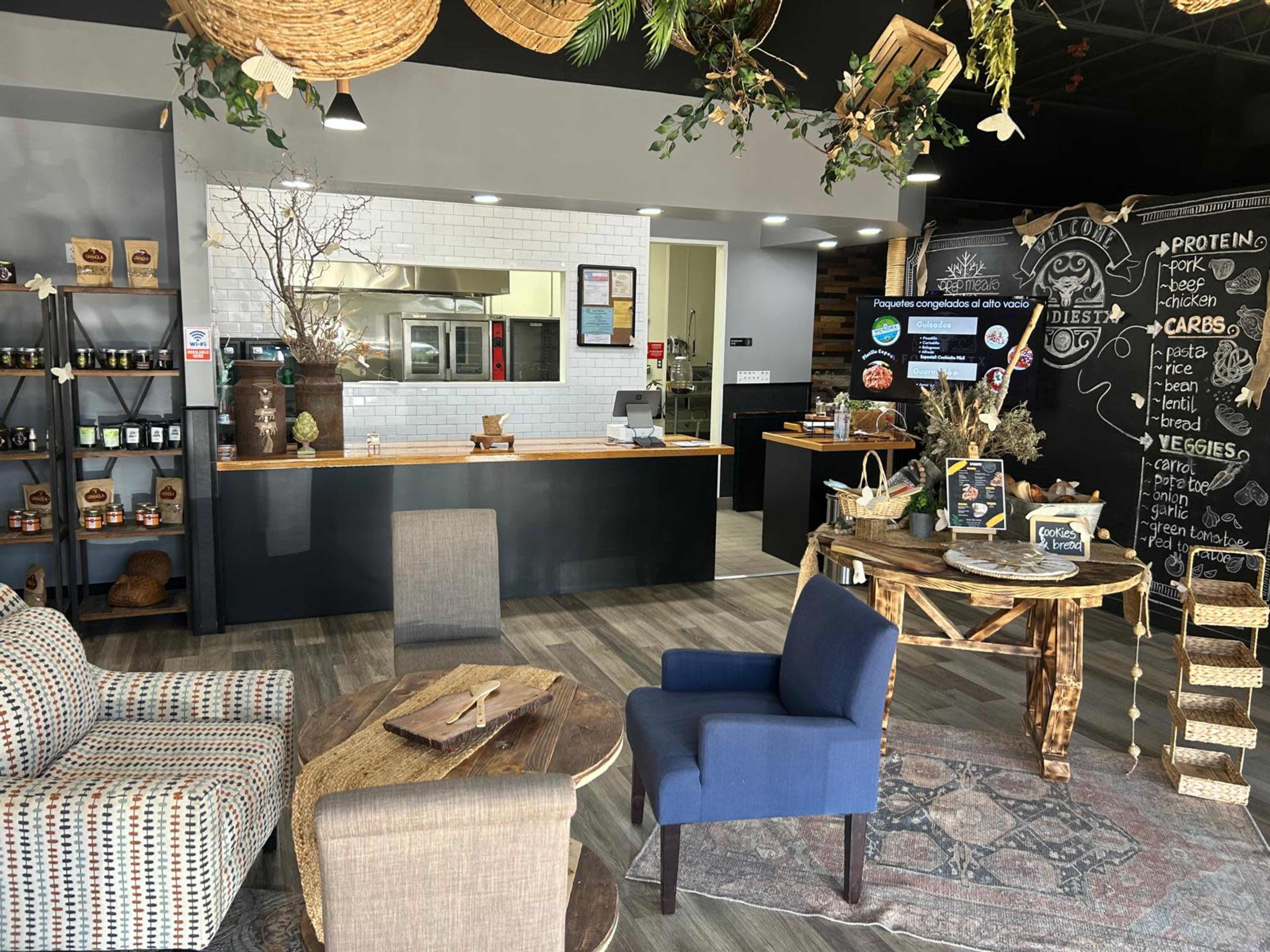 The image depicts a cozy café interior featuring a kitchen area, a seating arrangement with armchairs, and a chalkboard menu.