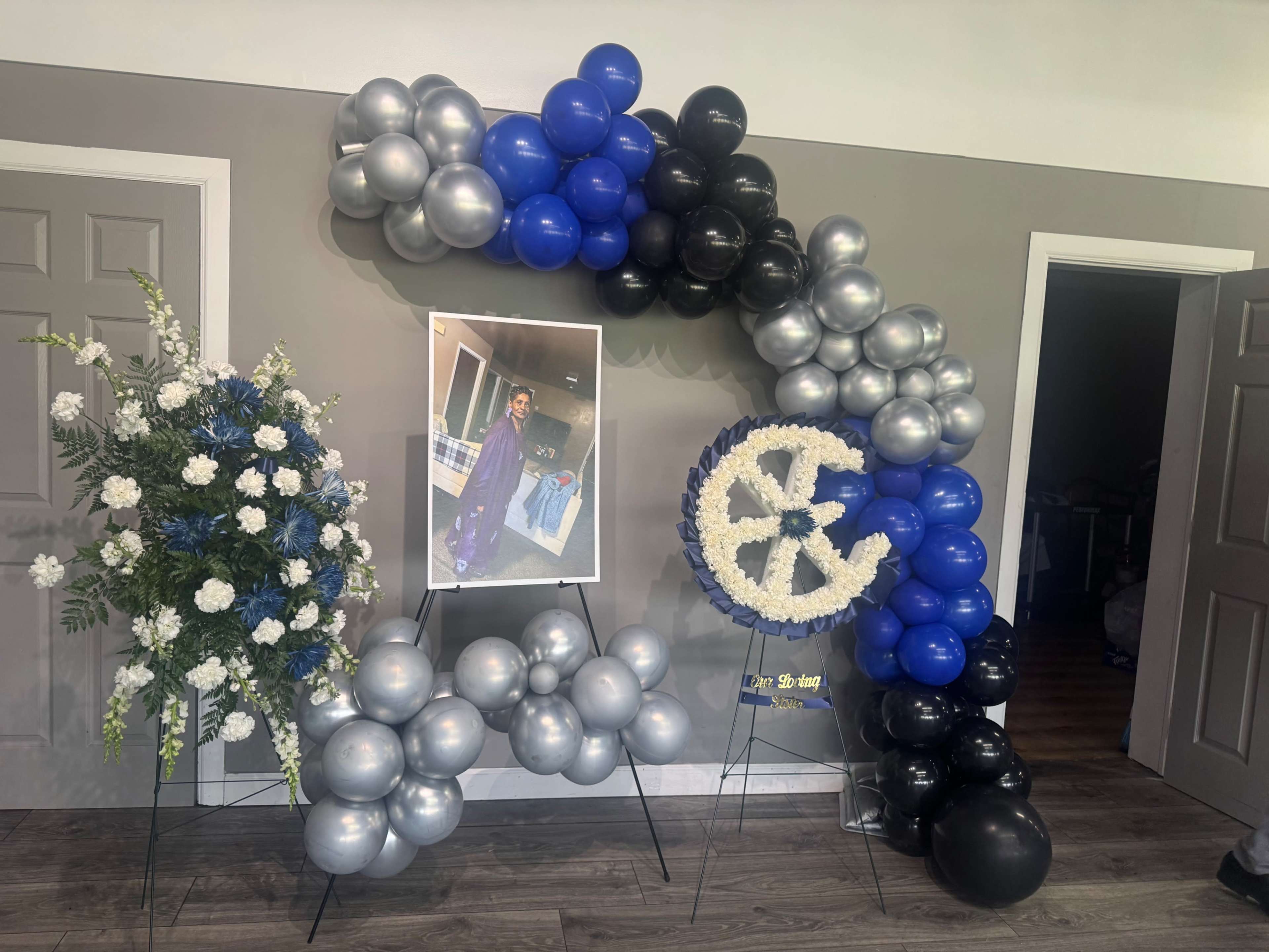 The image features a decorative arch made of blue, silver, and black balloons, alongside a floral arrangement and a framed photo displayed on easels.