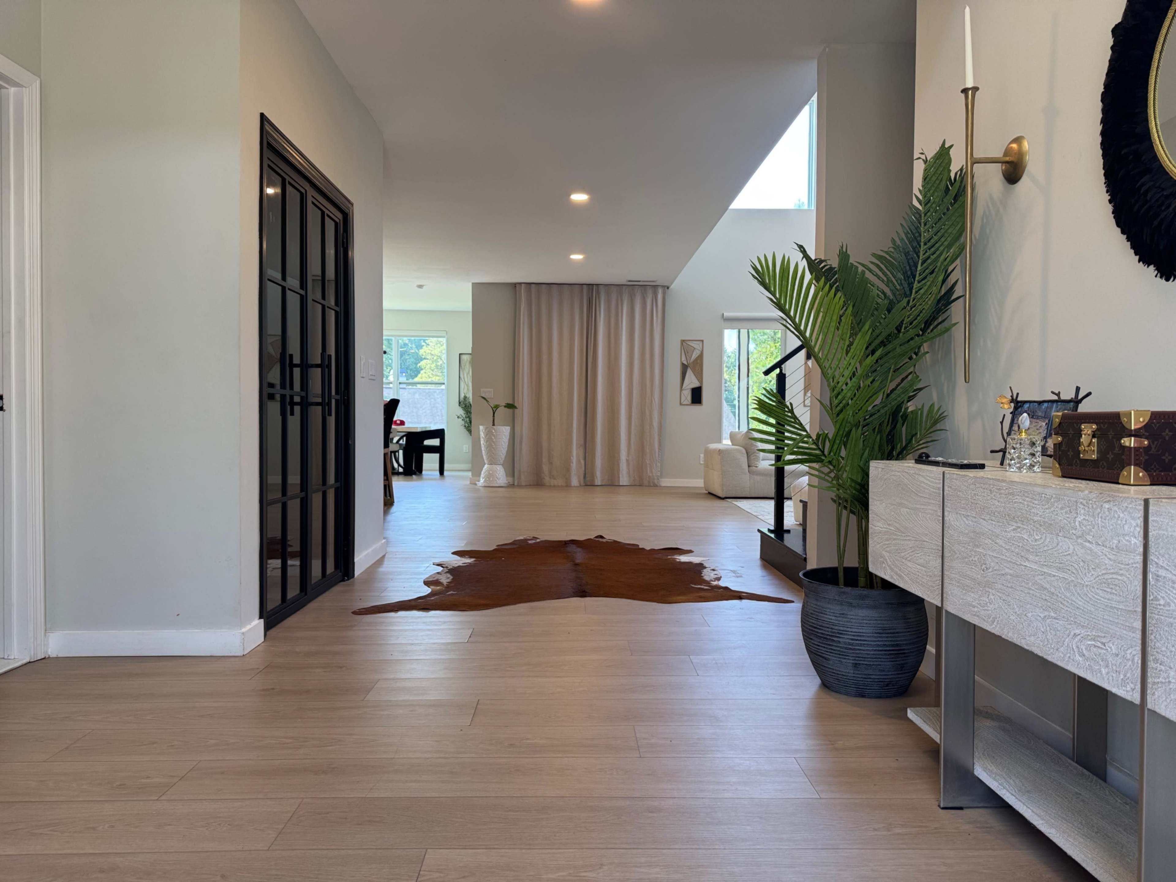 The image shows a spacious hallway leading to an open living area, featuring a decorative plant, a cowhide rug, and modern furniture.