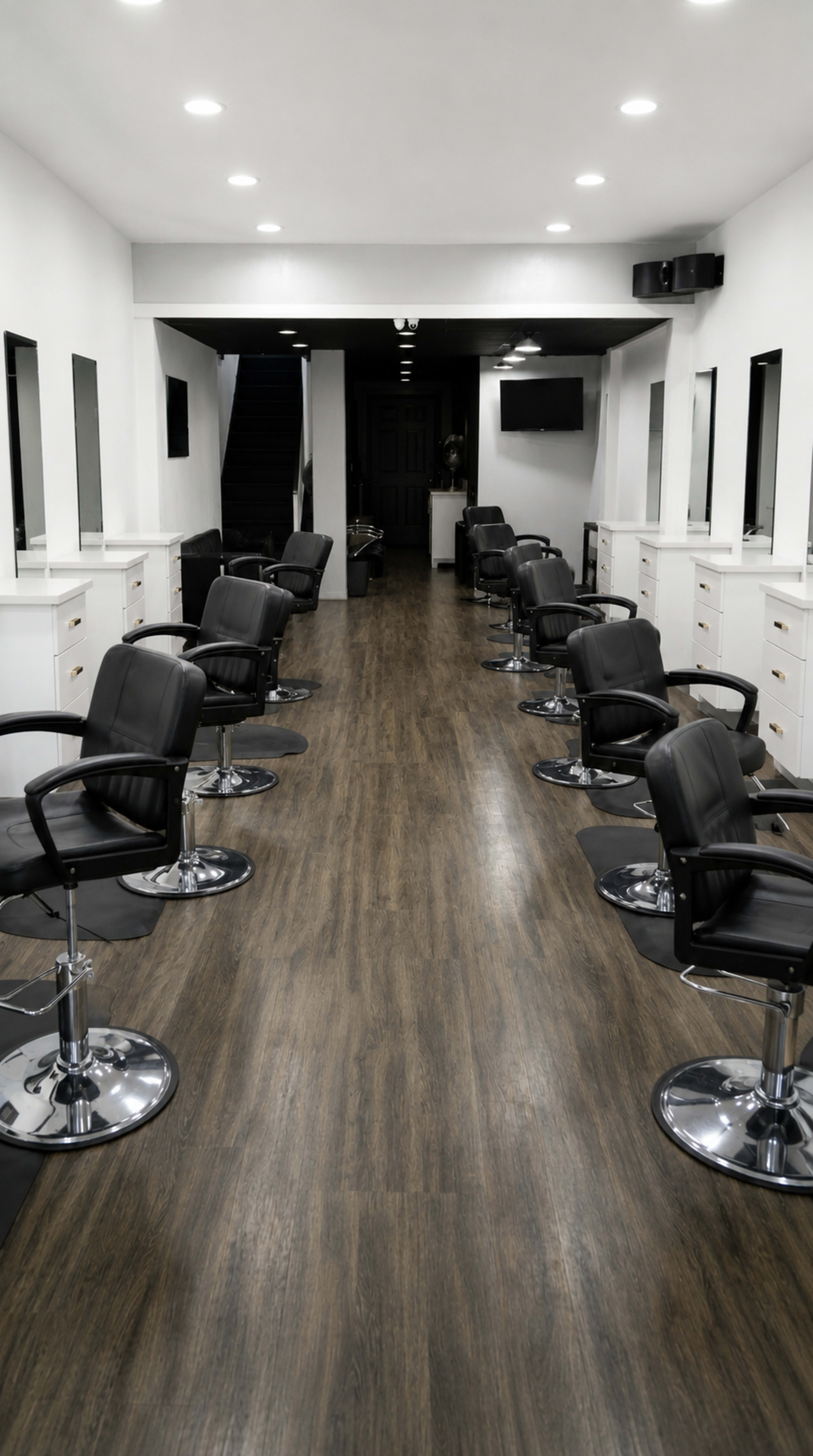 The image shows a modern hair salon with several black salon chairs arranged along a wooden floor and large mirrors on the walls.