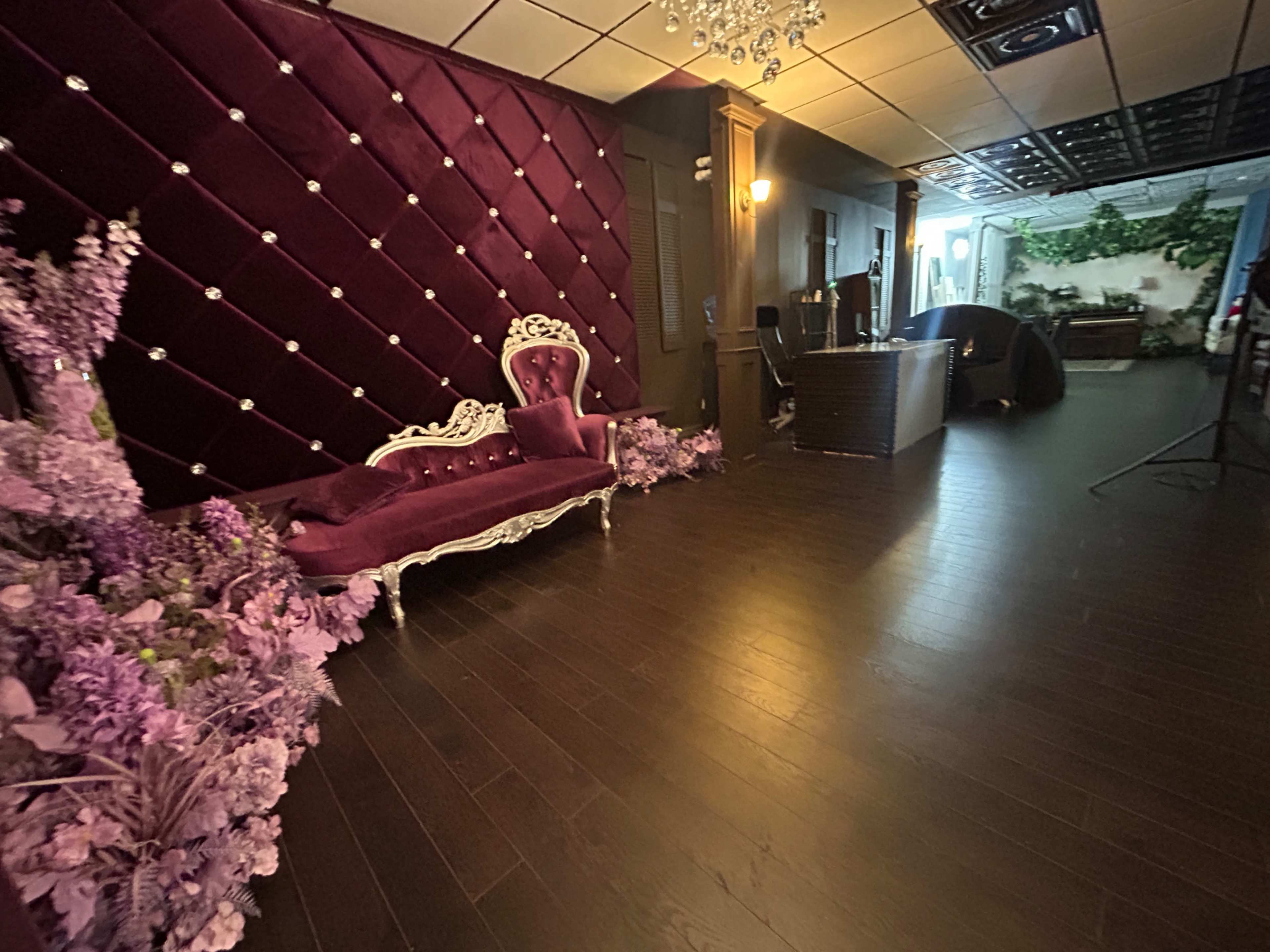 The image shows an elegantly decorated interior featuring a plush purple seating area with a vintage-style sofa, adorned with flowers and a chandelier.