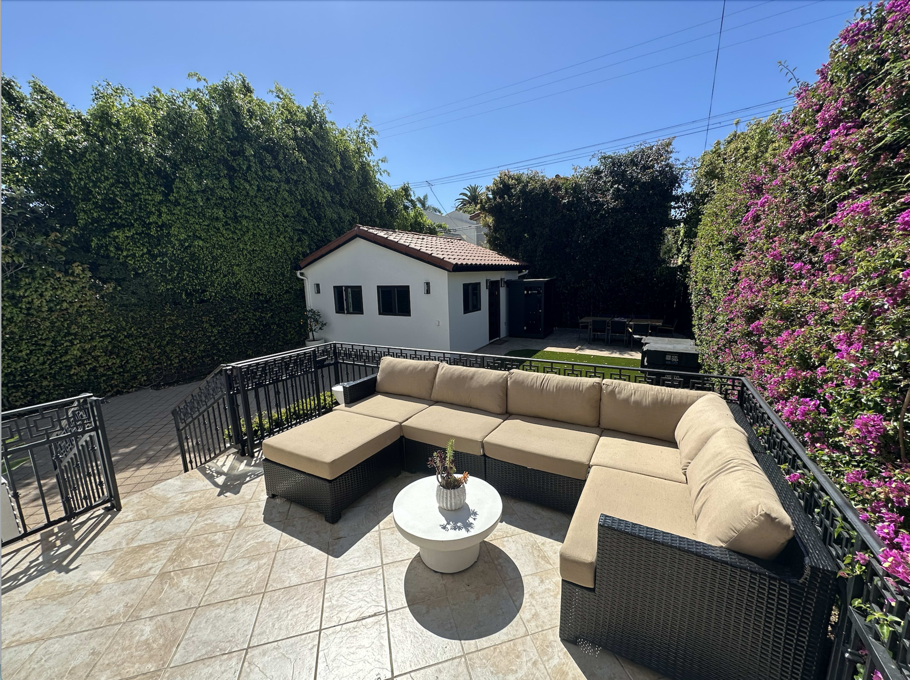 A patio area features a sectional sofa and a round table, surrounded by lush greenery and flowering plants, with a small house nearby.