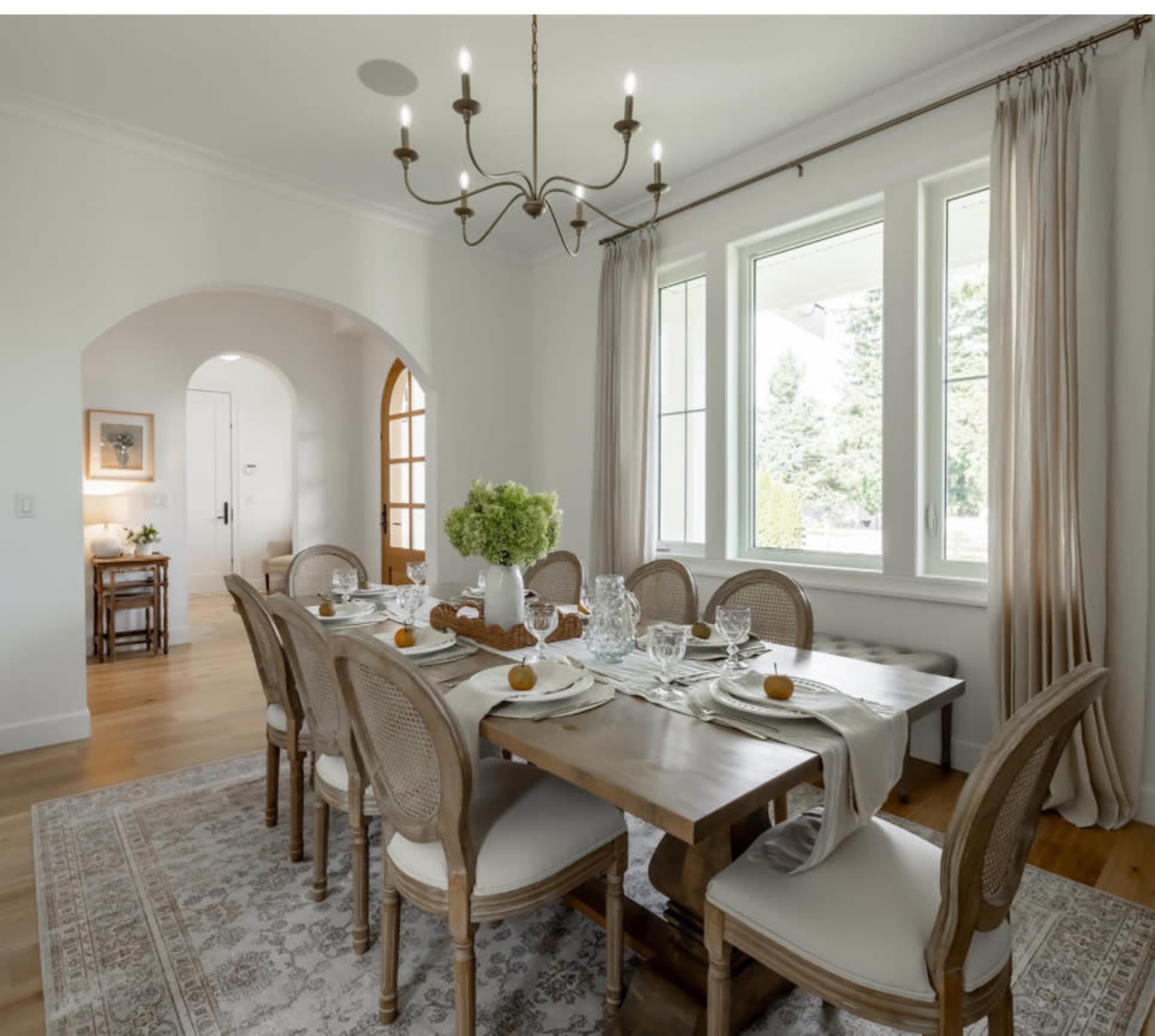 The image shows a well-decorated dining room with a large wooden table set for a meal, surrounded by eight chairs, and featuring large windows with natural light.