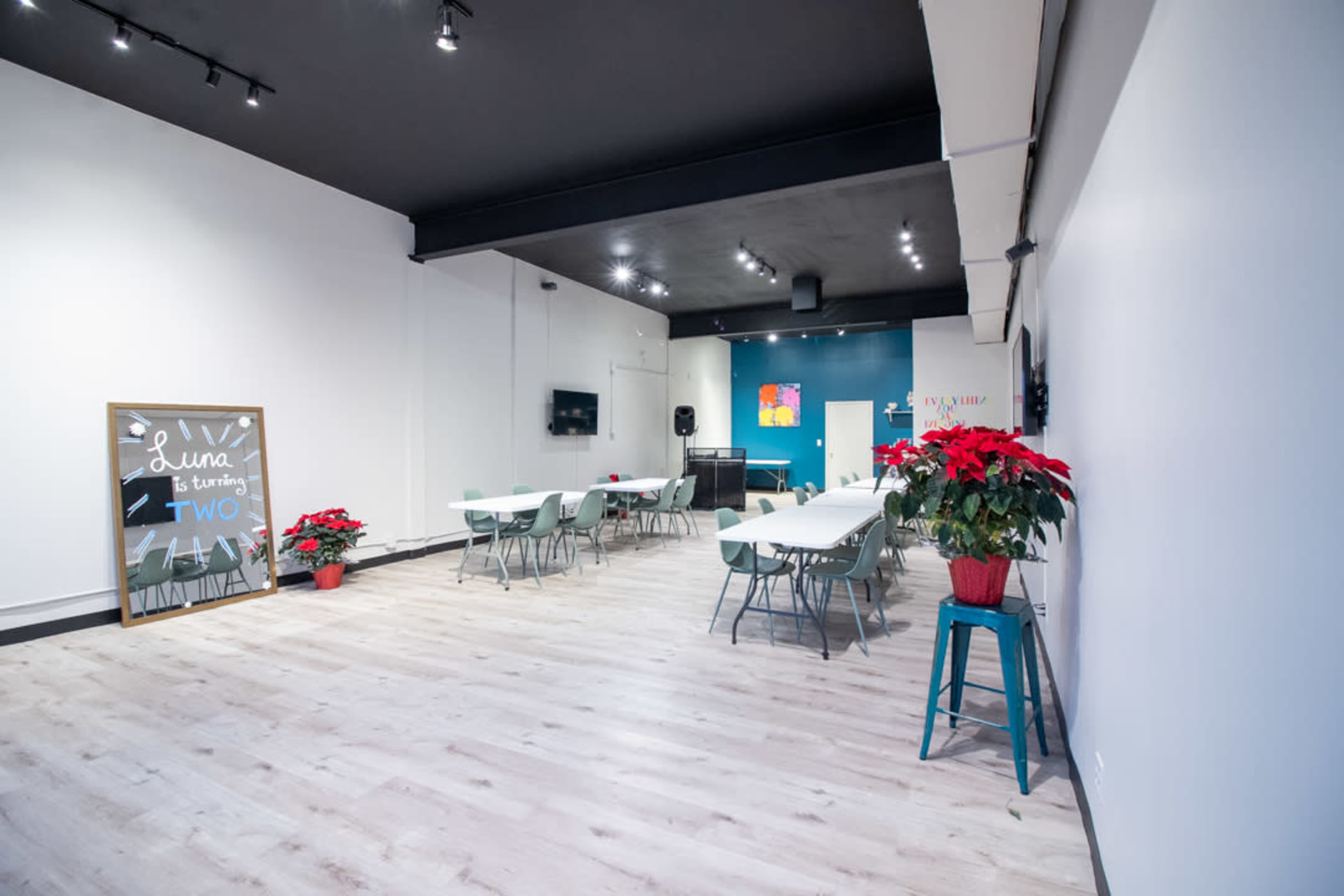 A spacious event room with tables and chairs arranged, a television on the wall, and a decorative sign announcing a birthday celebration, complemented by poinsettia plants.