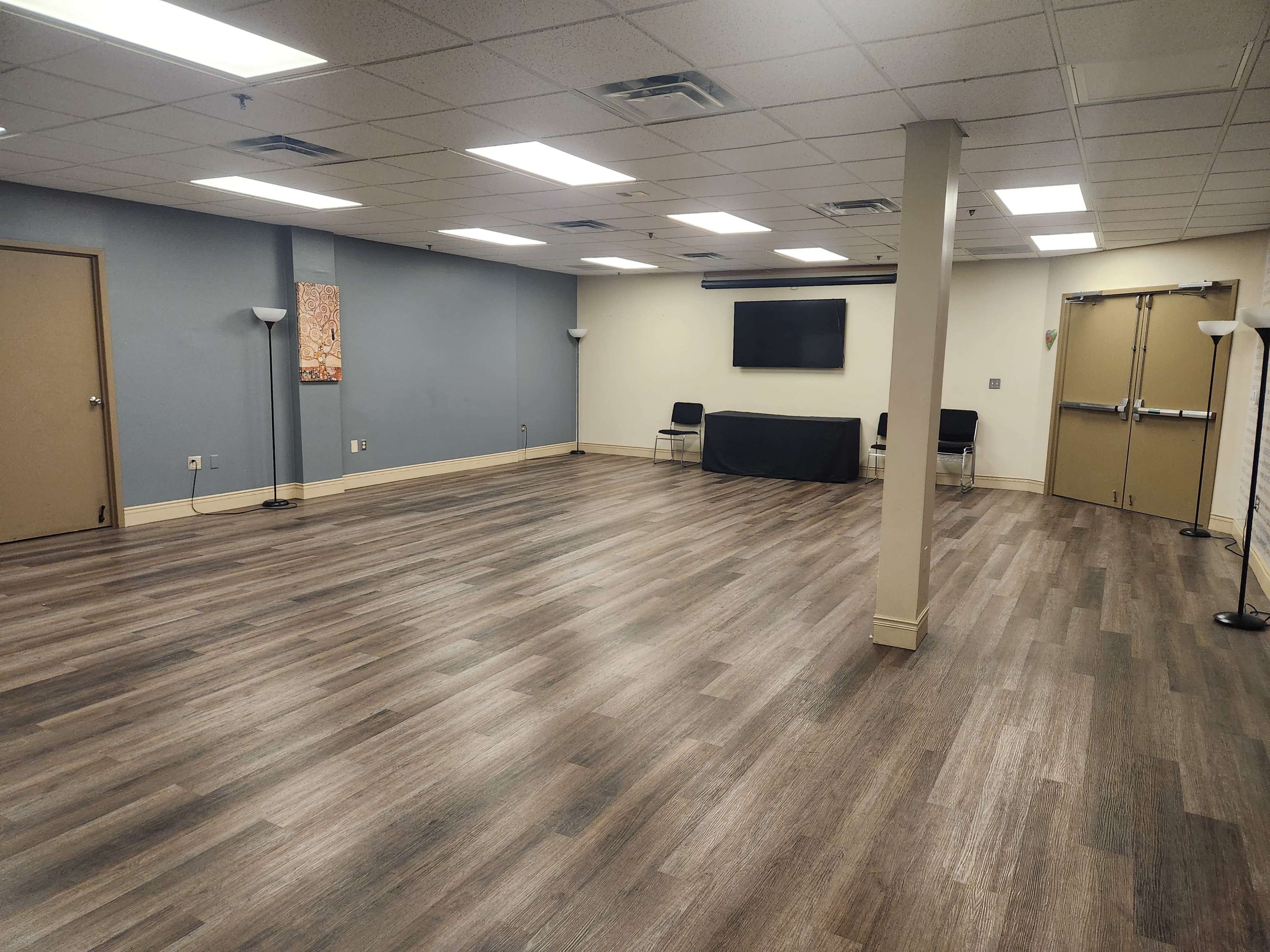 The image shows an empty room with hardwood flooring, fluorescent lighting, a flat-screen TV mounted on the wall, and a table with chairs in the center.