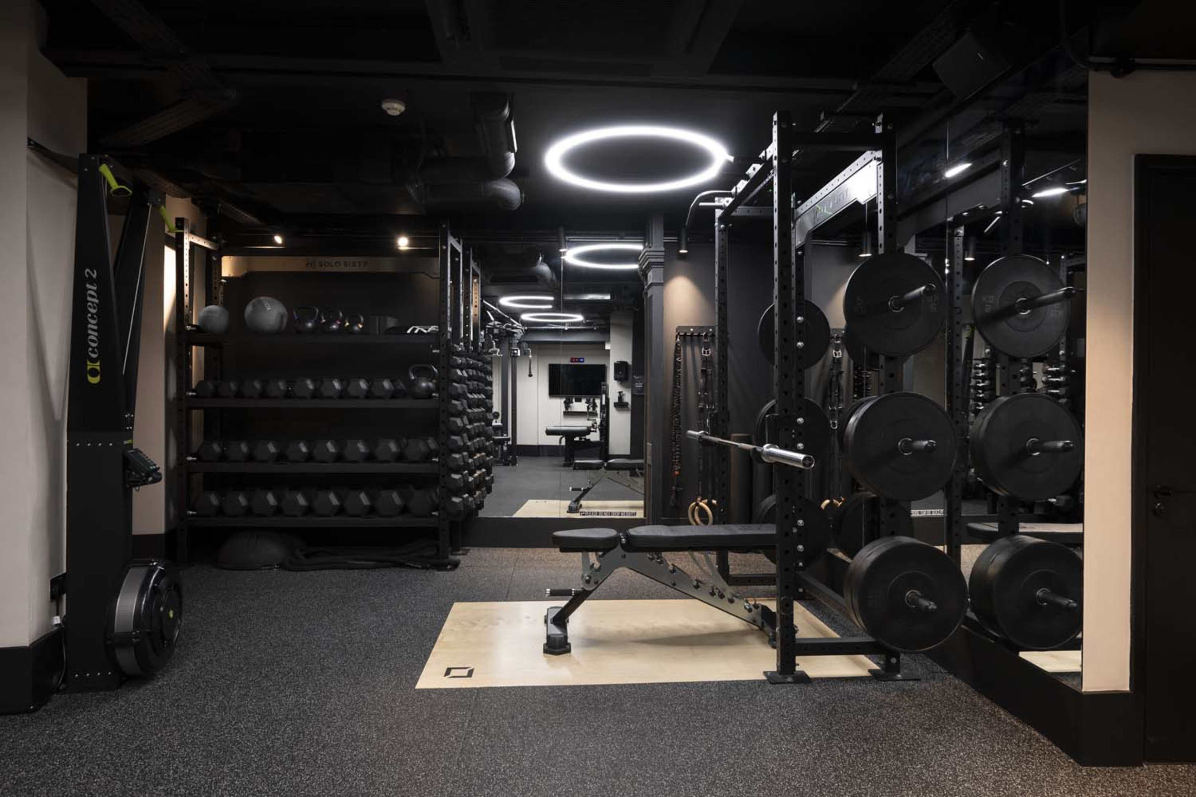 The image shows a modern gym with various weightlifting equipment, including benches, dumbbells, and resistance machines, illuminated by circular ceiling lights.