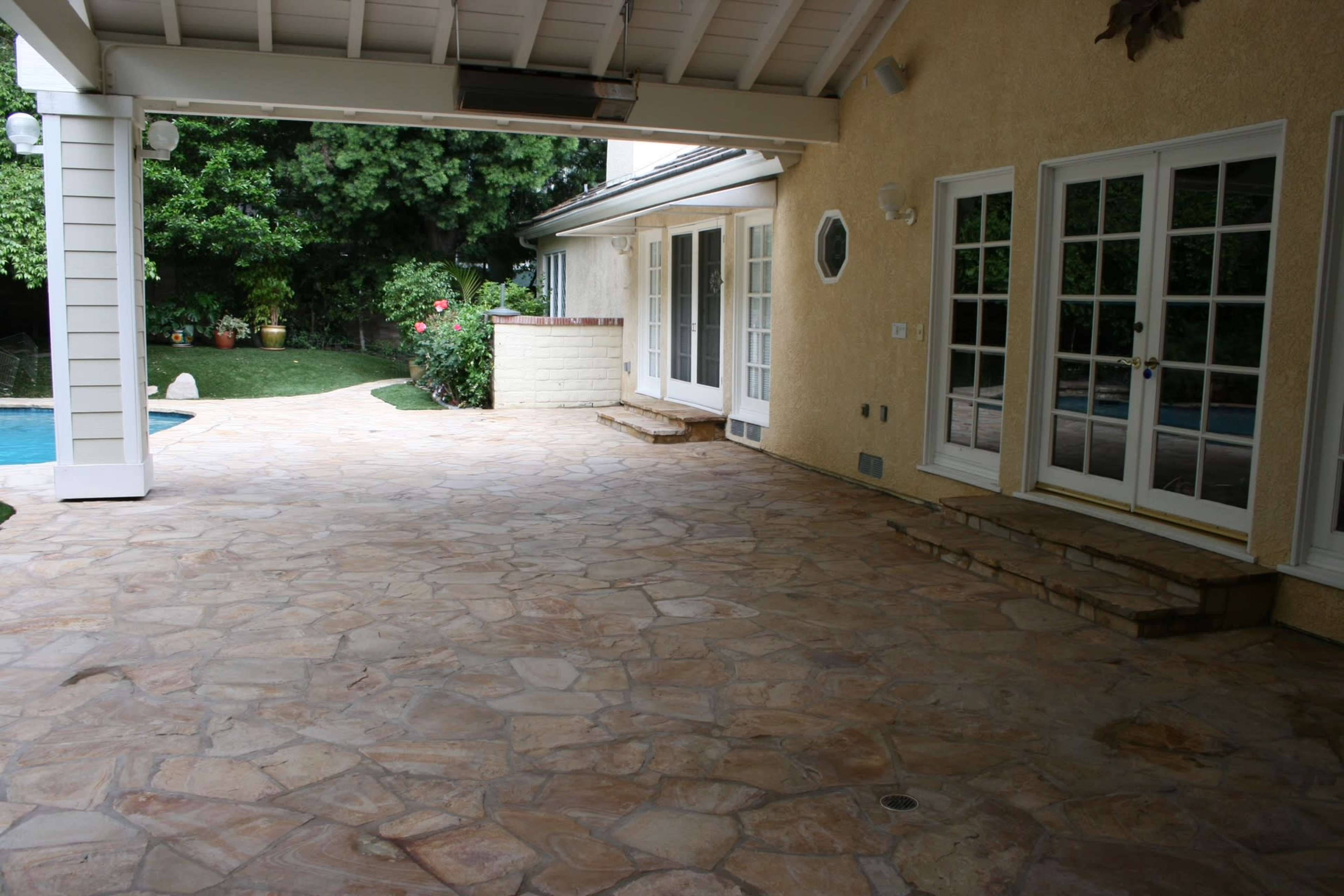 The image shows a covered outdoor patio with a stone floor, leading to a house with multiple French doors and a swimming pool visible in the background.