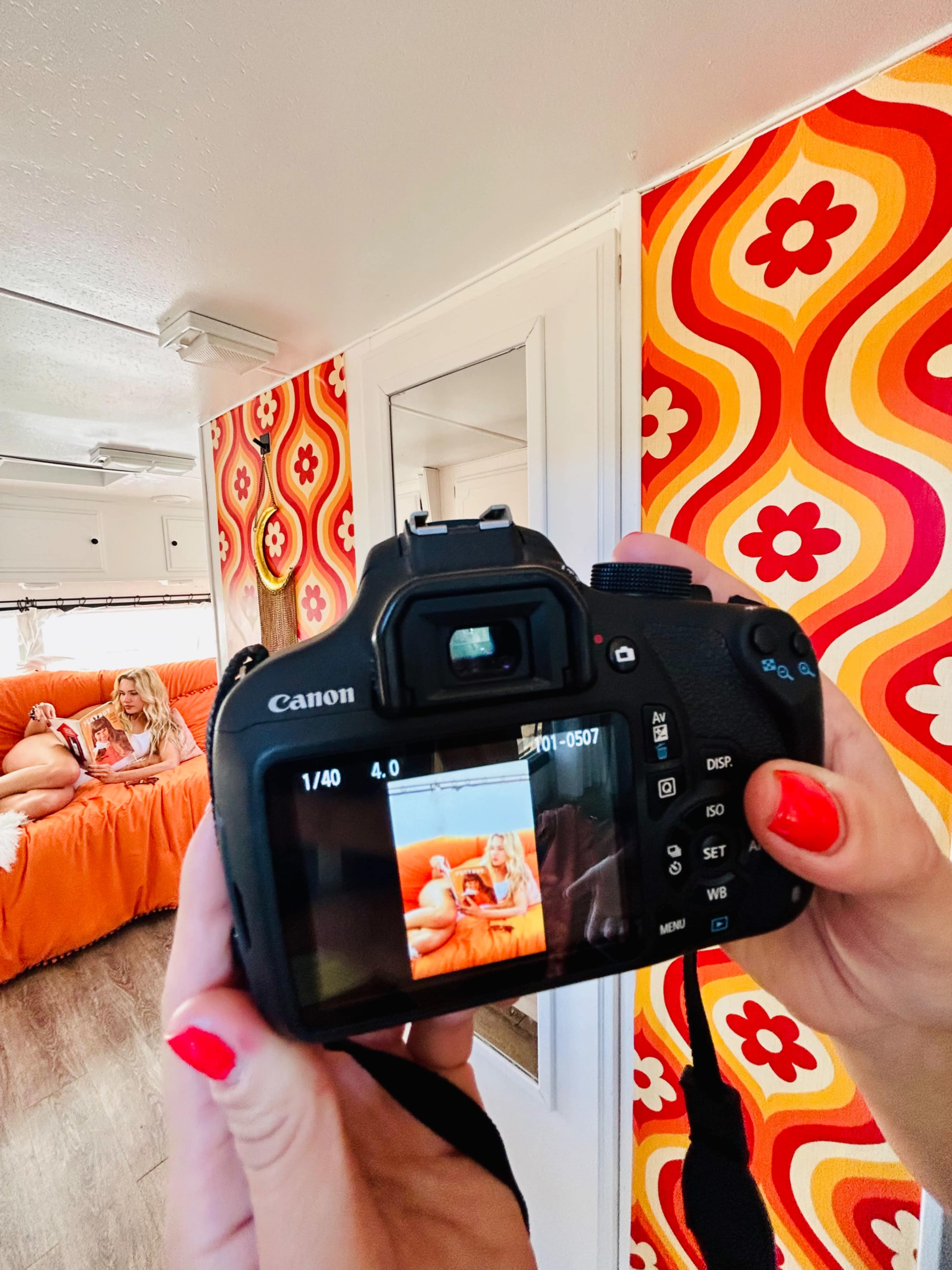 A person holds a Canon camera displaying a photo of a woman lounging on an orange couch against a vibrant, retro-style wall.