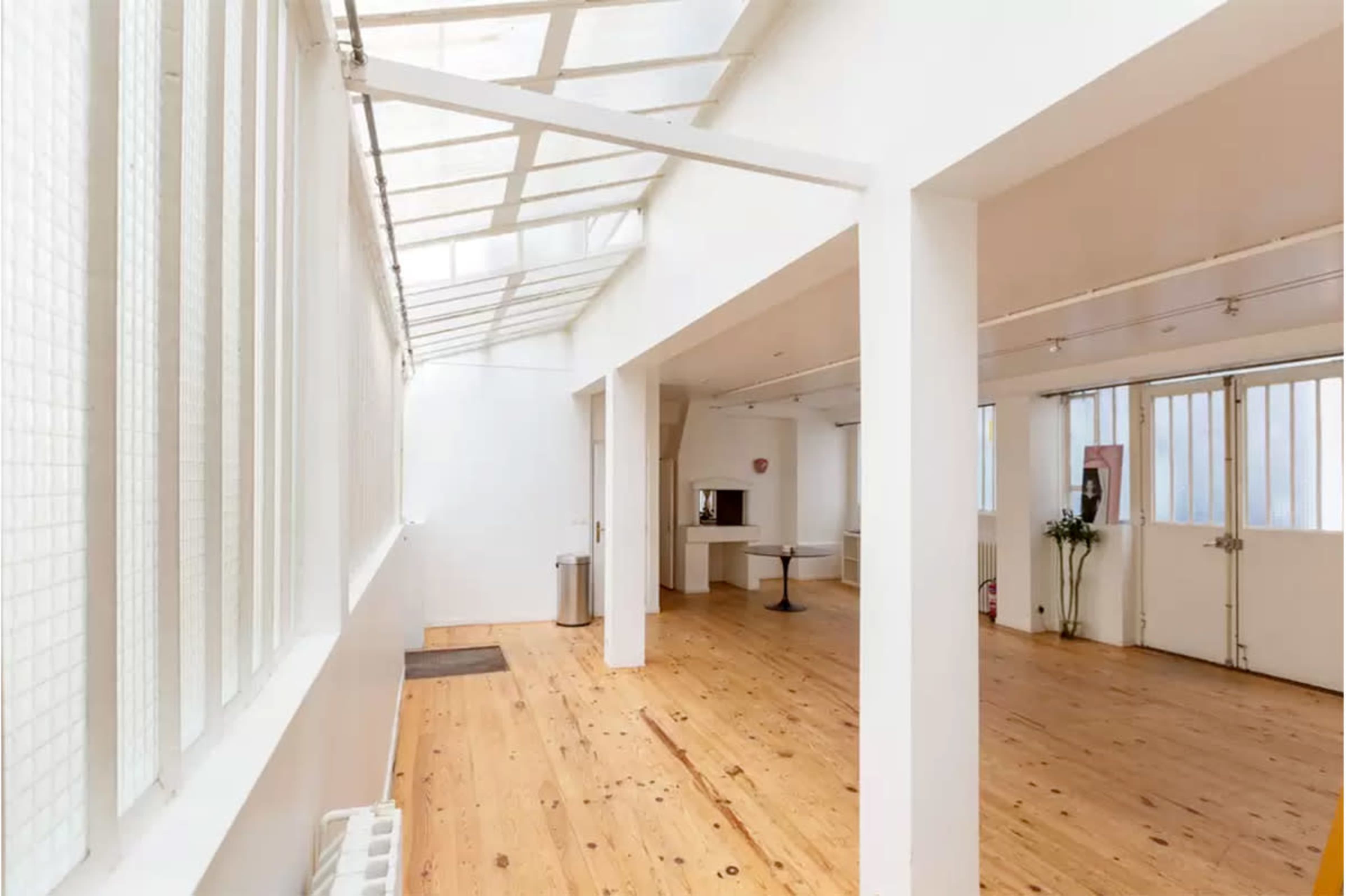A bright, spacious room with wooden flooring, large windows, and minimal furnishings.